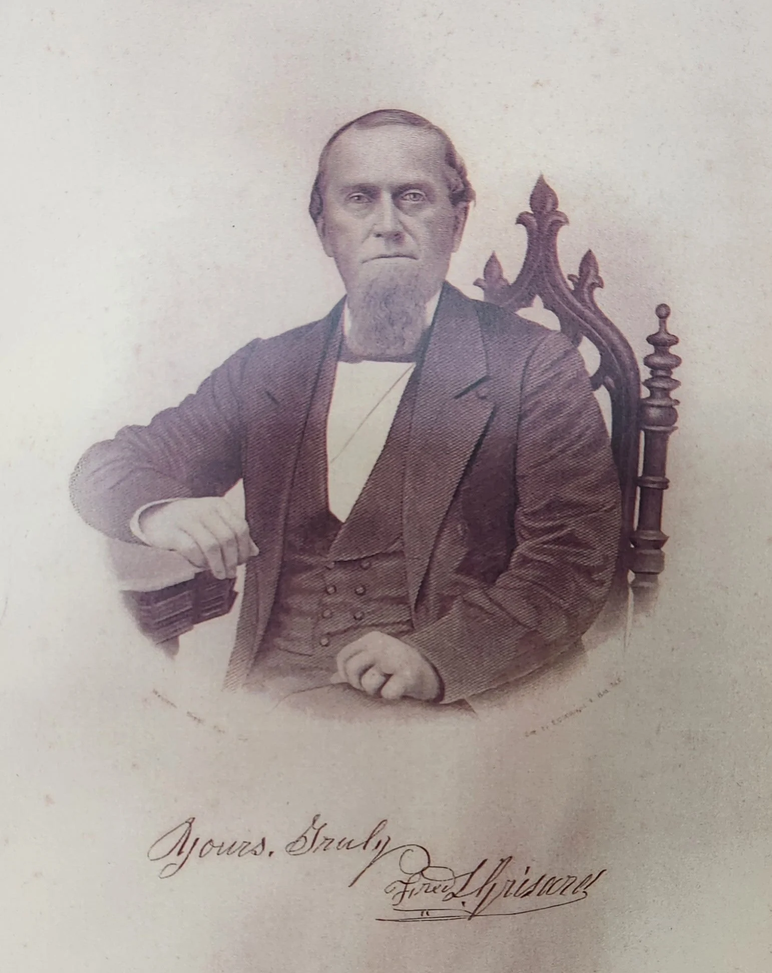 A black and white historical portrait of an older man with a beard sitting in a wooden high-backed chair, dressed in 19th-century clothing. There is a handwritten inscription below the portrait.