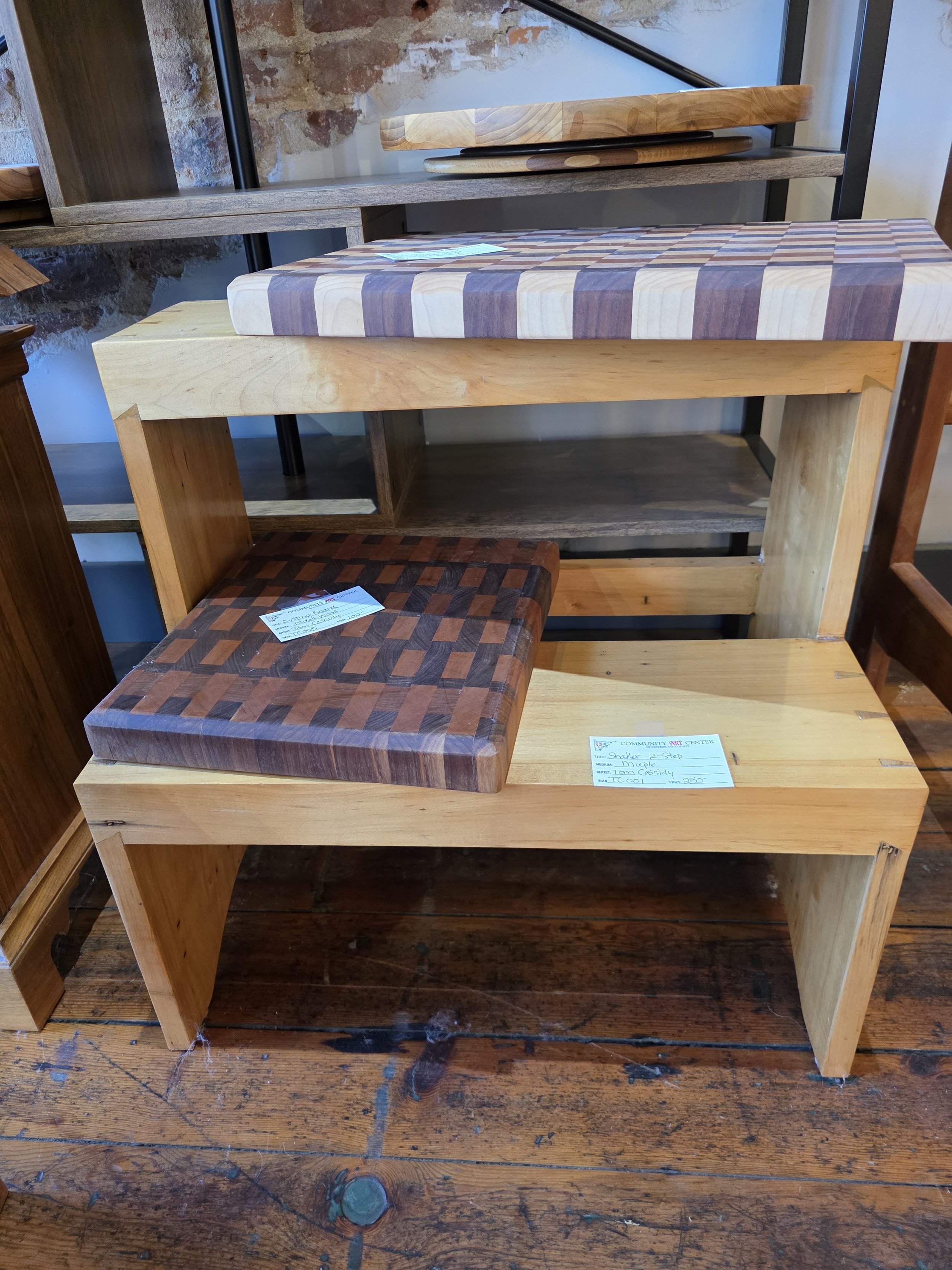 A wooden bench with a square, checkered seat made of different wood shades, displayed in a furniture store. There is a price tag on the seat and another card on the right side of the bench.