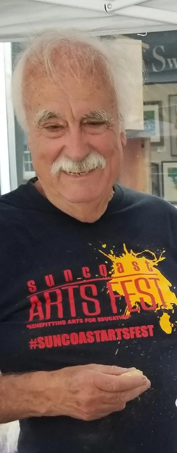 An older man with white hair and a mustache smiling, wearing a black T-shirt with red and yellow text for the Suncoast Arts Fest.