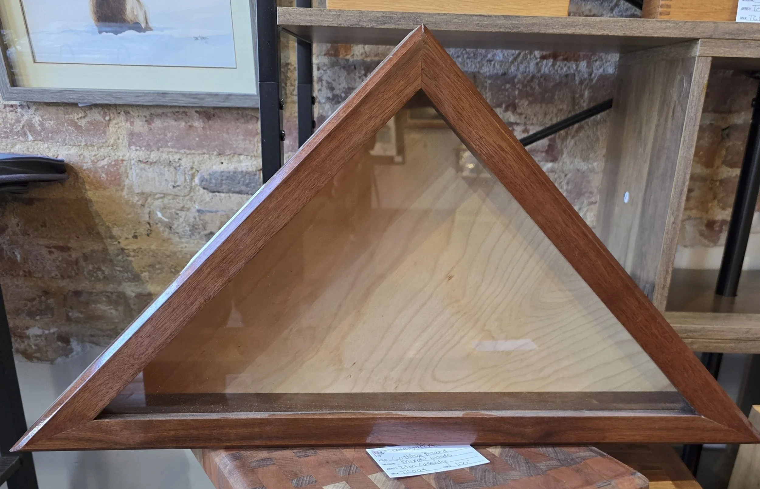 A triangular wooden and glass display case on a wooden surface in front of a brick wall, with a framed picture hanging on the wall and a wooden furniture piece in the background.