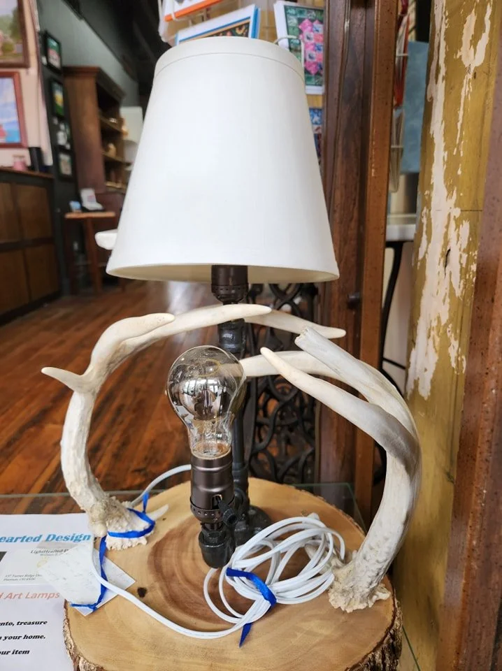 A decorative table lamp featuring antler-like arms and a light bulb at the center, with a white lampshade on top. The lamp is placed on a wooden surface in a room with wooden walls and various decorations.