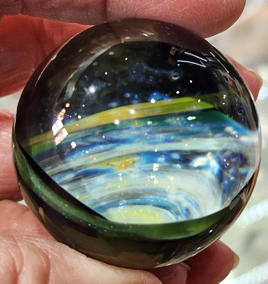 Close-up of a hand holding a glass marble with a swirl of blue, green, yellow, and white colors inside, reflecting light.