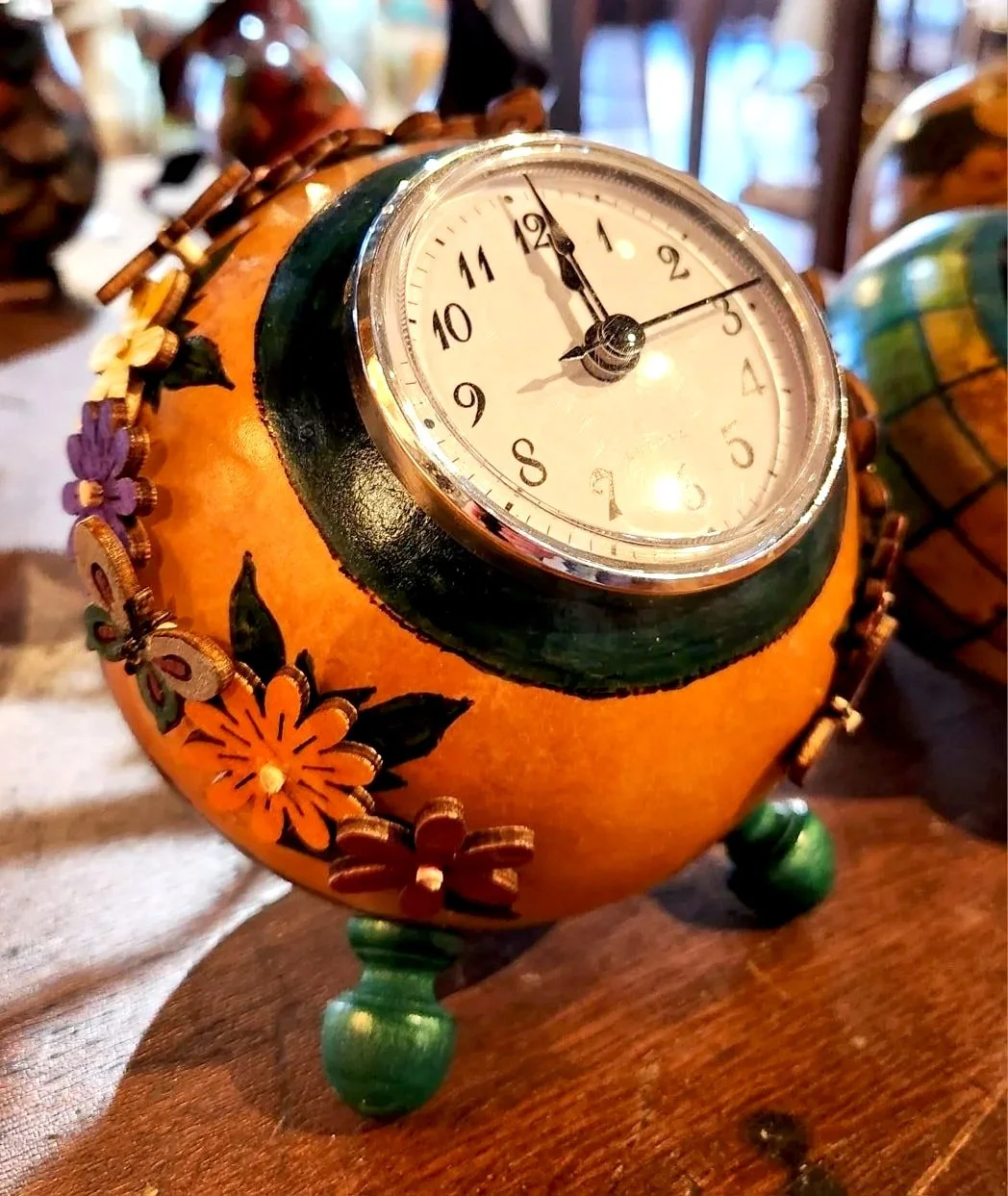 Decorative clock embedded on a round, orange gourd with floral designs, standing on three small green legs.