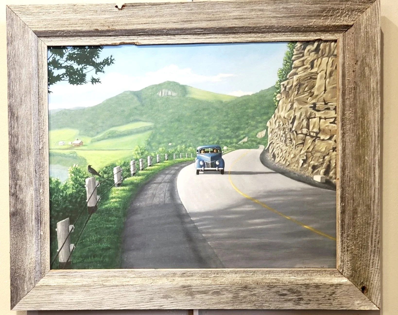 A painting of a winding country road with a vintage blue car driving along it, surrounded by lush green hills and a mountain in the background, framed with a rustic wood border.