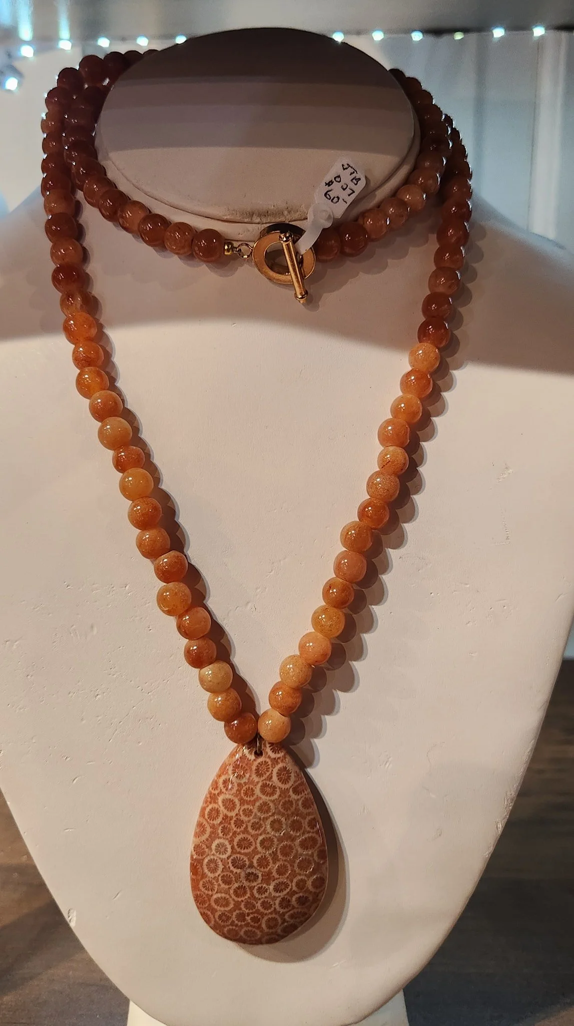 A necklace with peach-colored beads and a large teardrop-shaped pendant with a circular pattern, displayed on a white necklace stand.