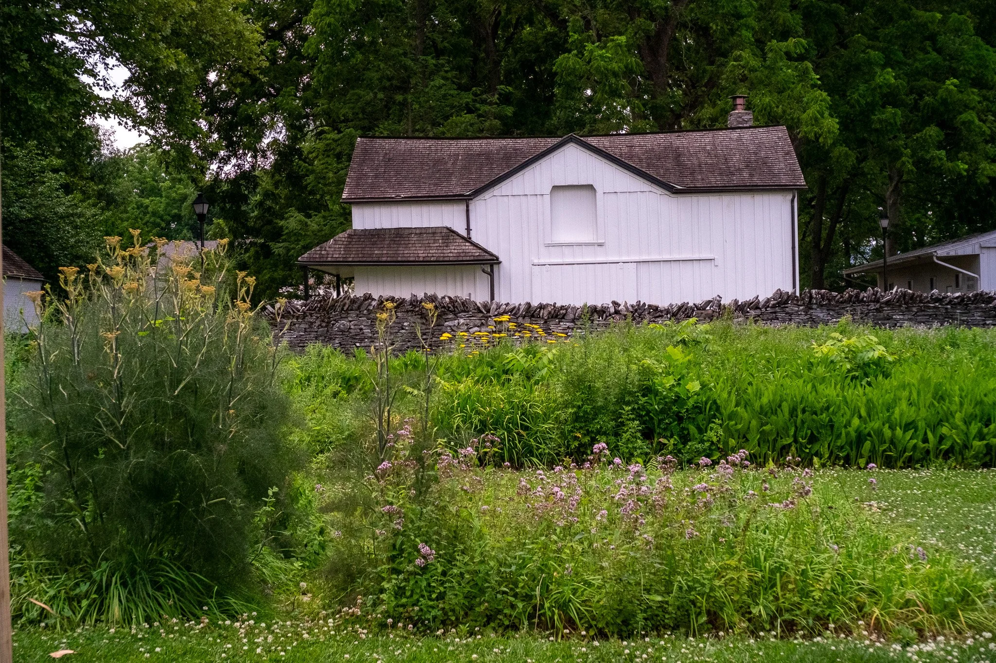 A white house with a shingled roof and a stone chimney, surrounded by lush green trees and a garden with flowering plants and a stone wall.