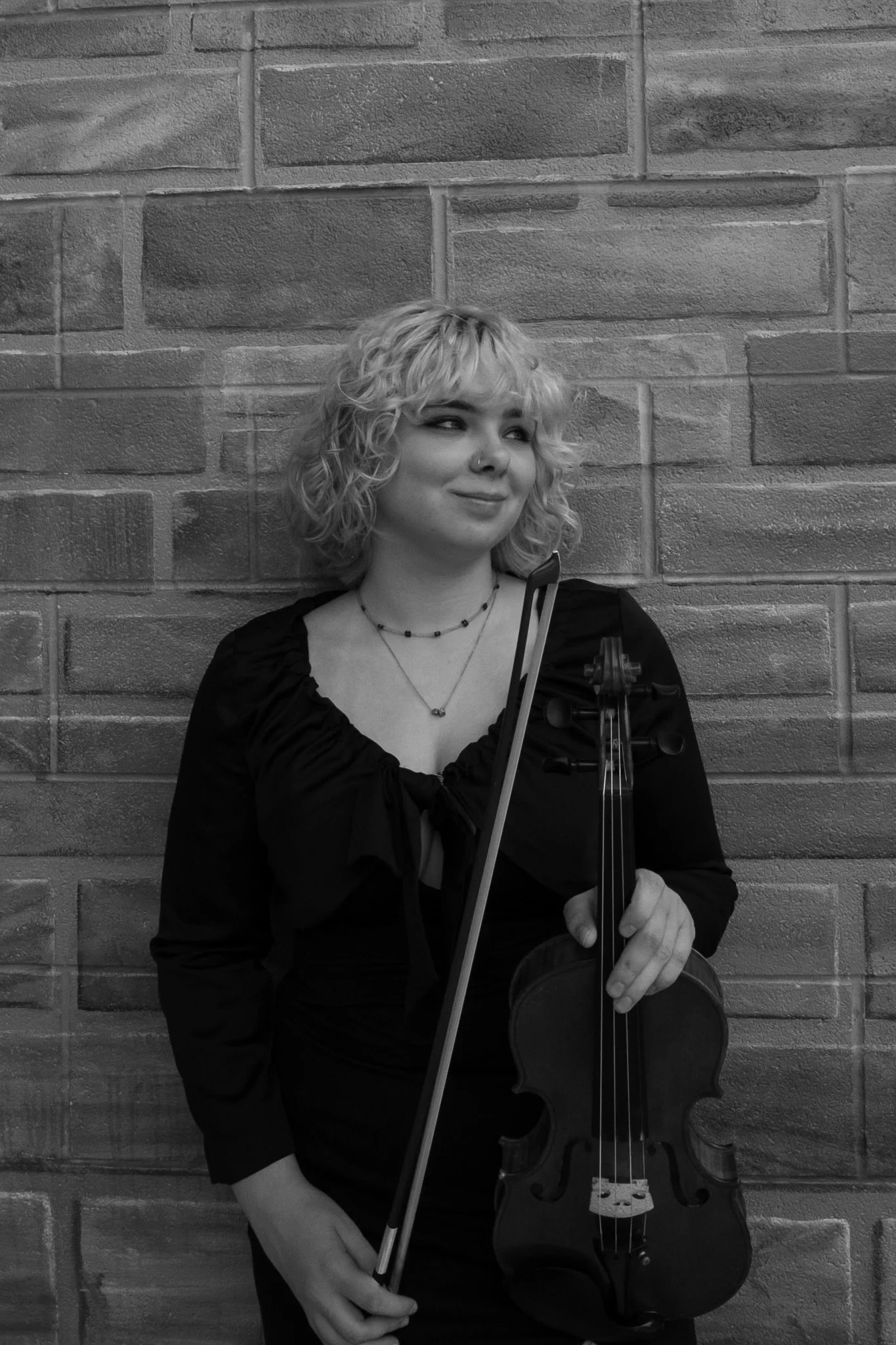 A young woman with curly blonde hair holding a violin and bow, standing against a brick wall, wearing a black dress and a necklace, looking to her right.