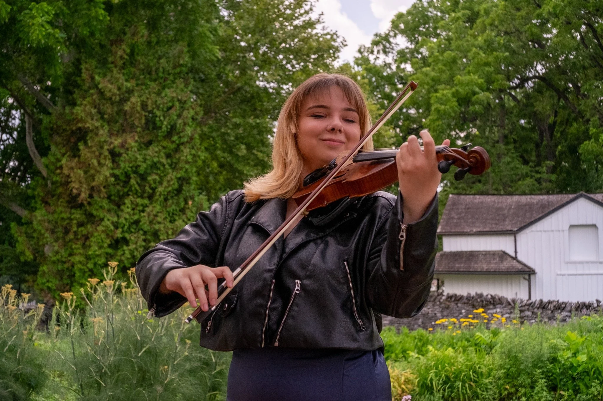 A woman playing the violin outdoors in a garden, with trees and a white building in the background.