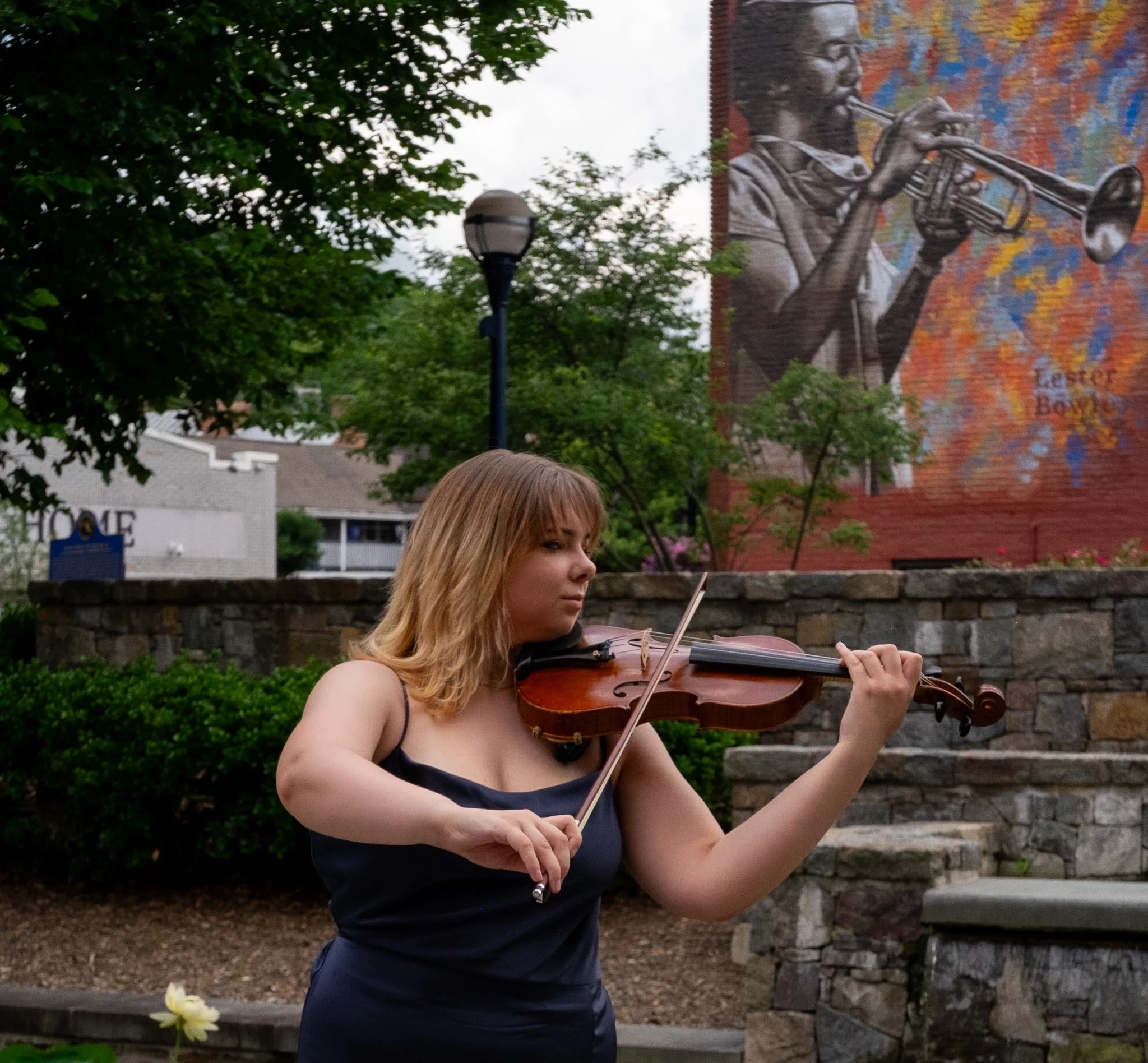 A woman with blonde hair playing a violin outdoors in front of trees and a brick wall with a colorful mural.