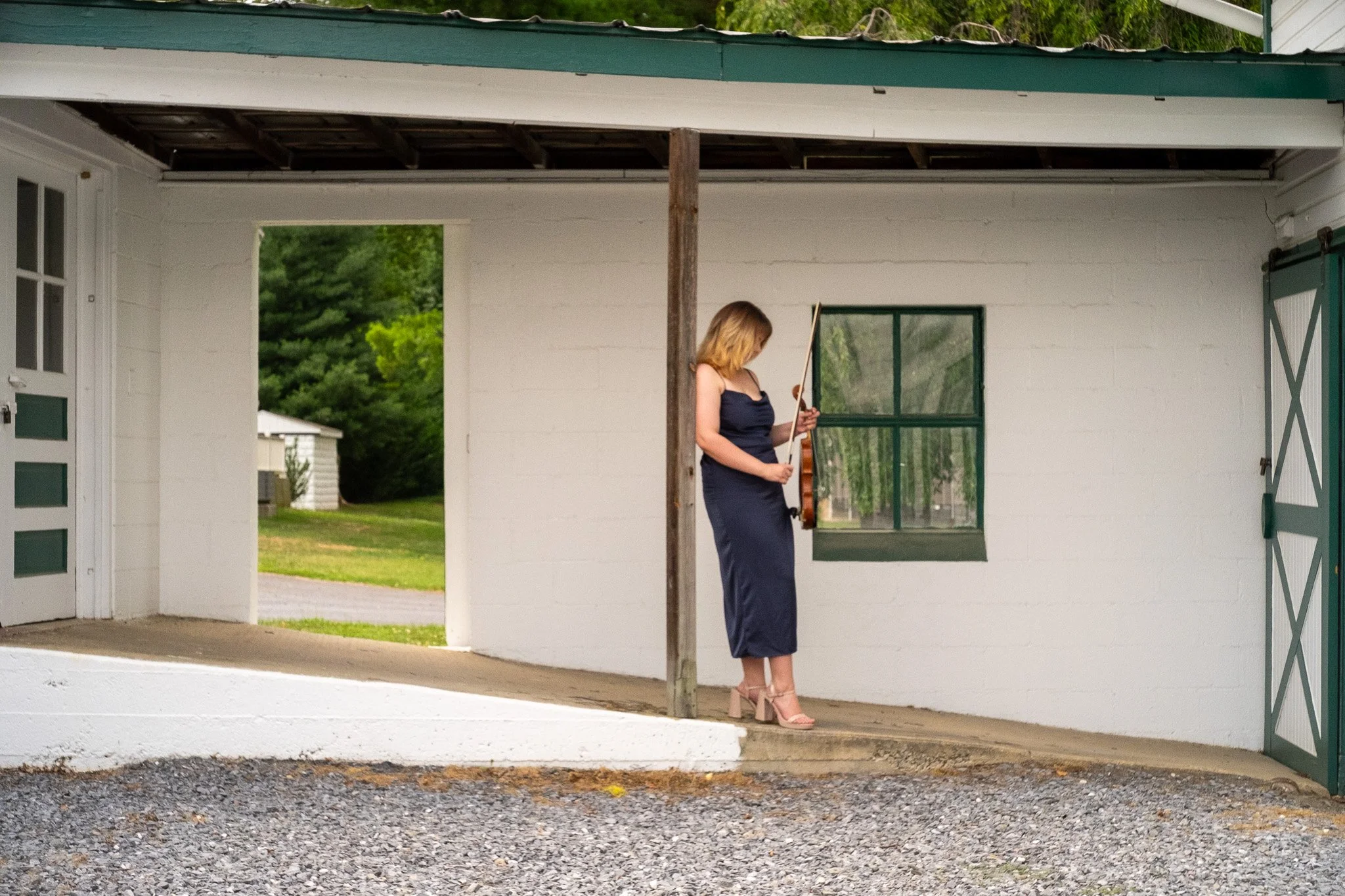 A woman in a long, elegant dark dress and high heels stands inside a garage, looking at a violin she is holding, with a background of green trees and a white house.