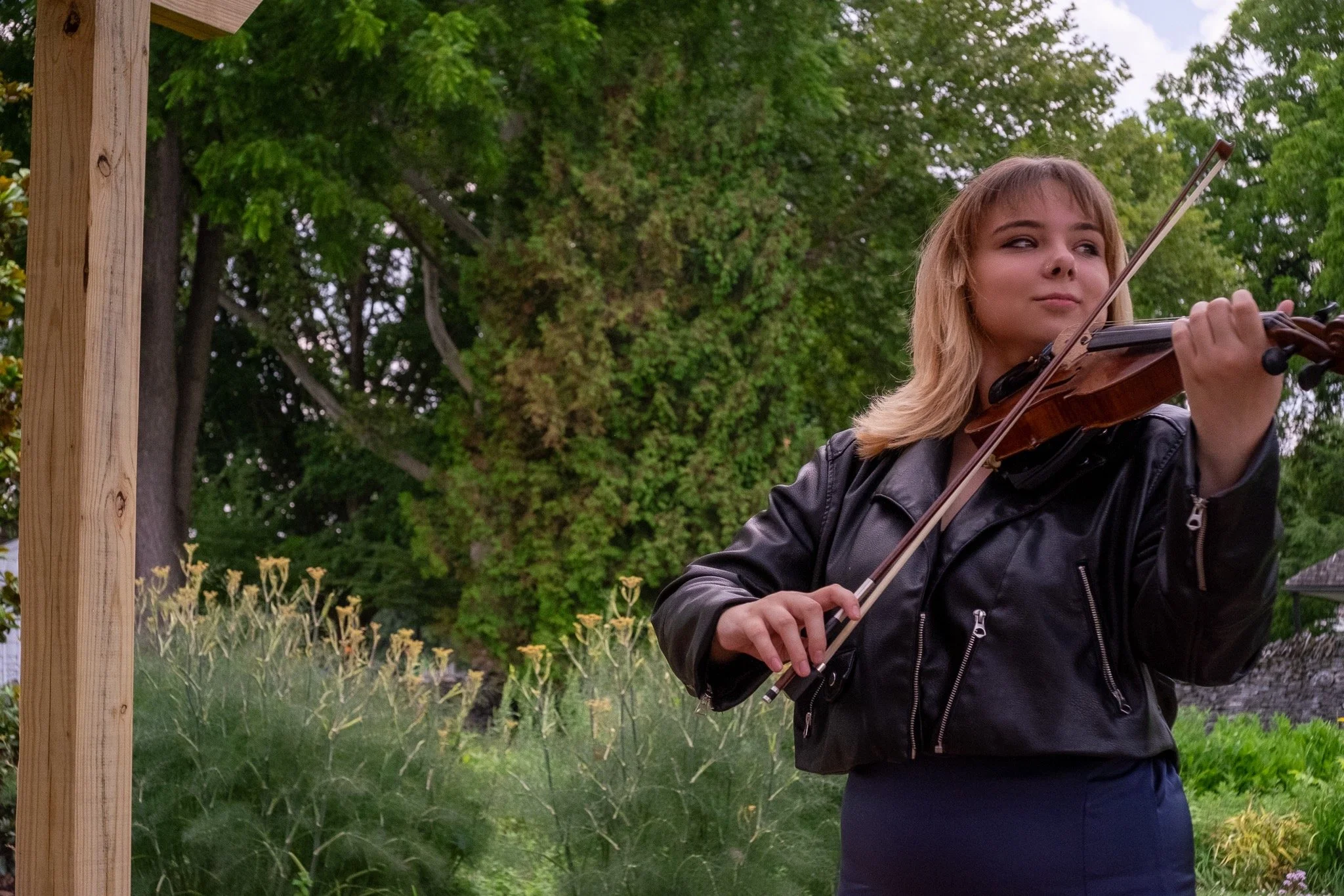 A young woman with blonde hair playing the violin outdoors near a wooden structure, with green trees and plants in the background.