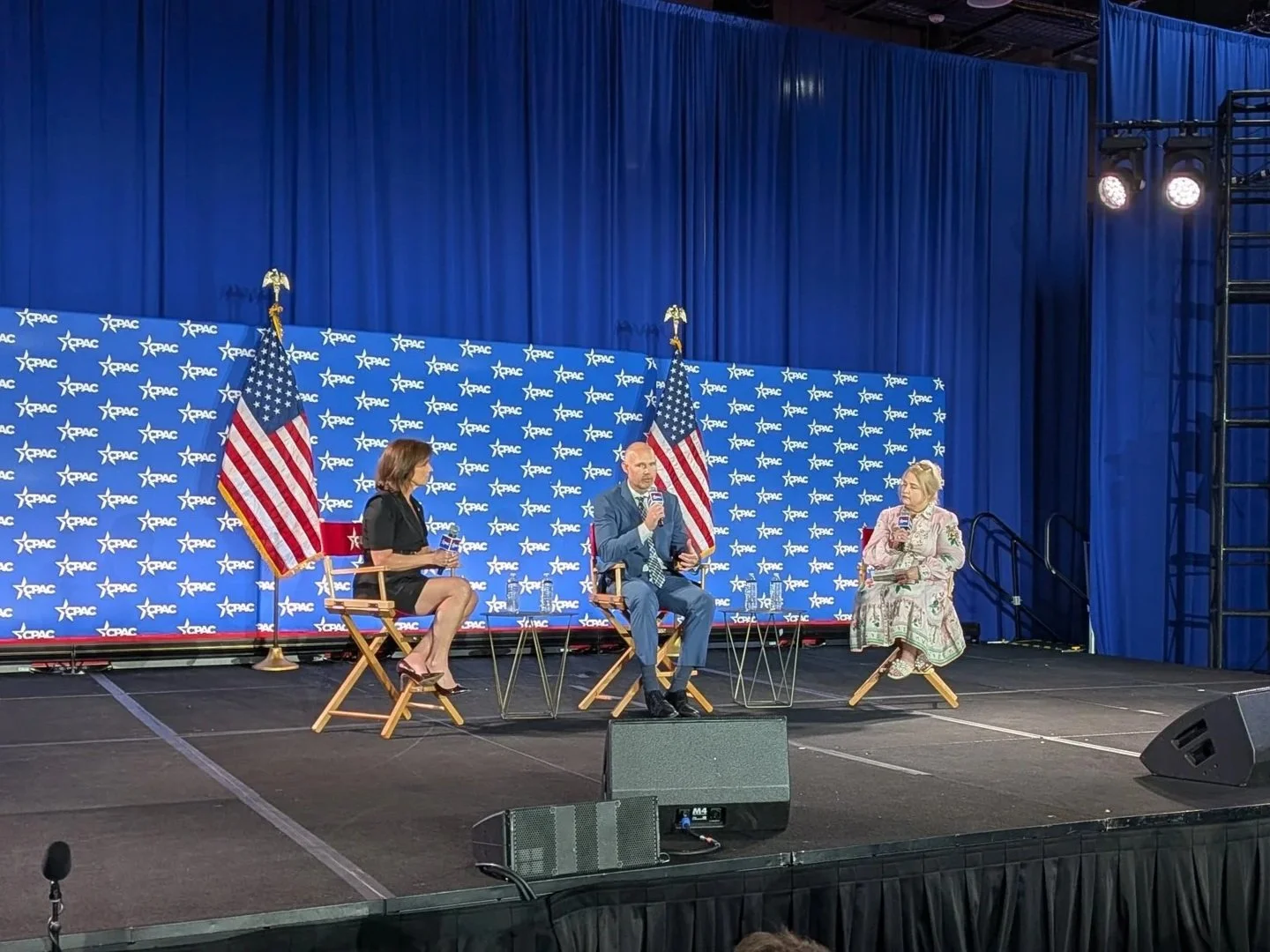 A panel discussion on stage at an event with a blue backdrop and U.S. flags. Three people are seated on chairs, two women and one man, with microphones.