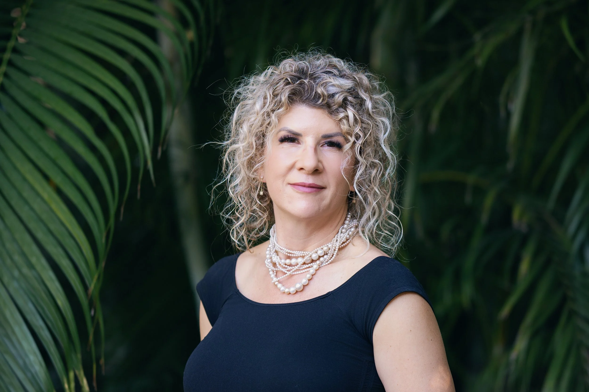 A woman with curly blonde hair posing outdoors in front of green foliage, wearing a black top and layered pearl necklaces.