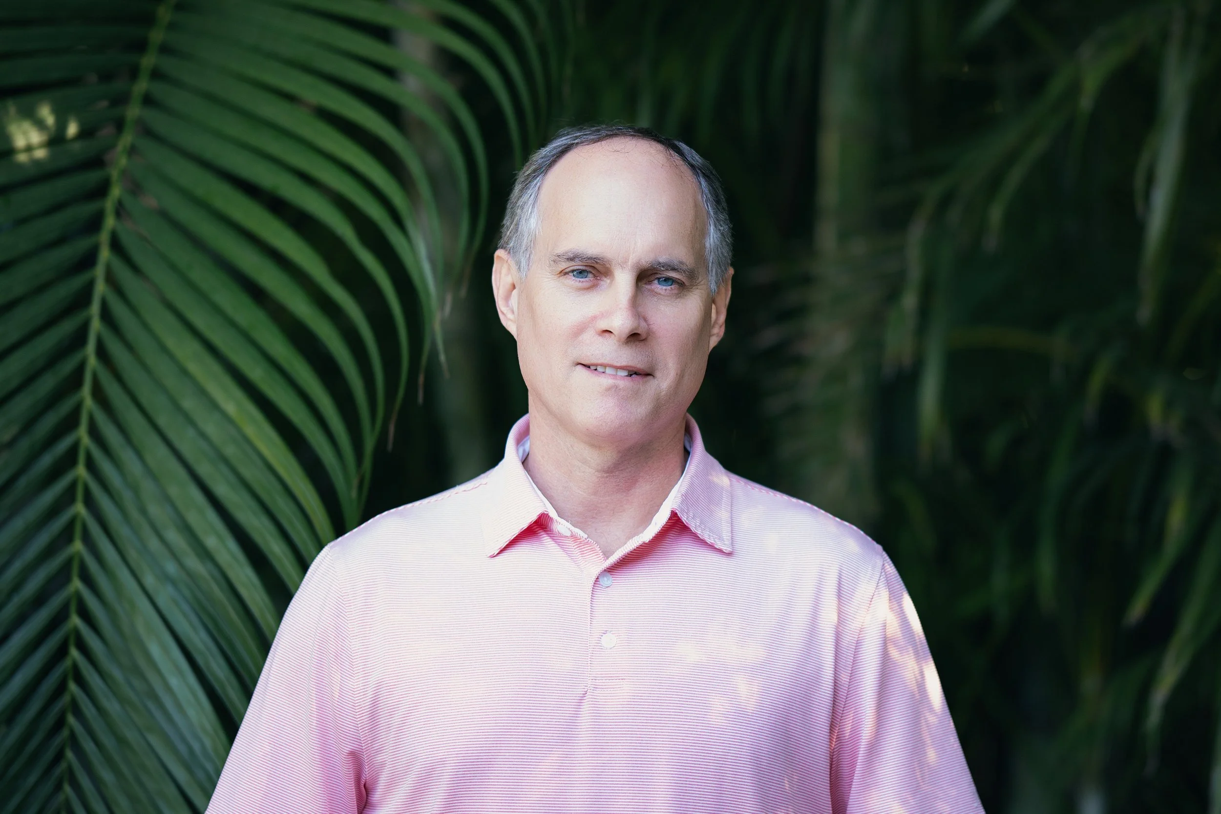 A middle-aged man with short gray hair and blue eyes stands outdoors in front of lush green palm leaves, wearing a light pink collared shirt.