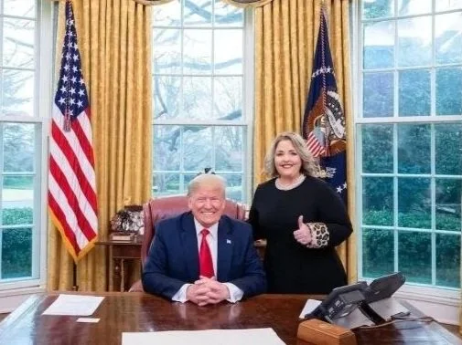Former President Donald Trump sitting at the White House desk with a woman standing beside him, both smiling and giving a thumbs-up. The background includes large windows with gold curtains and American flags.