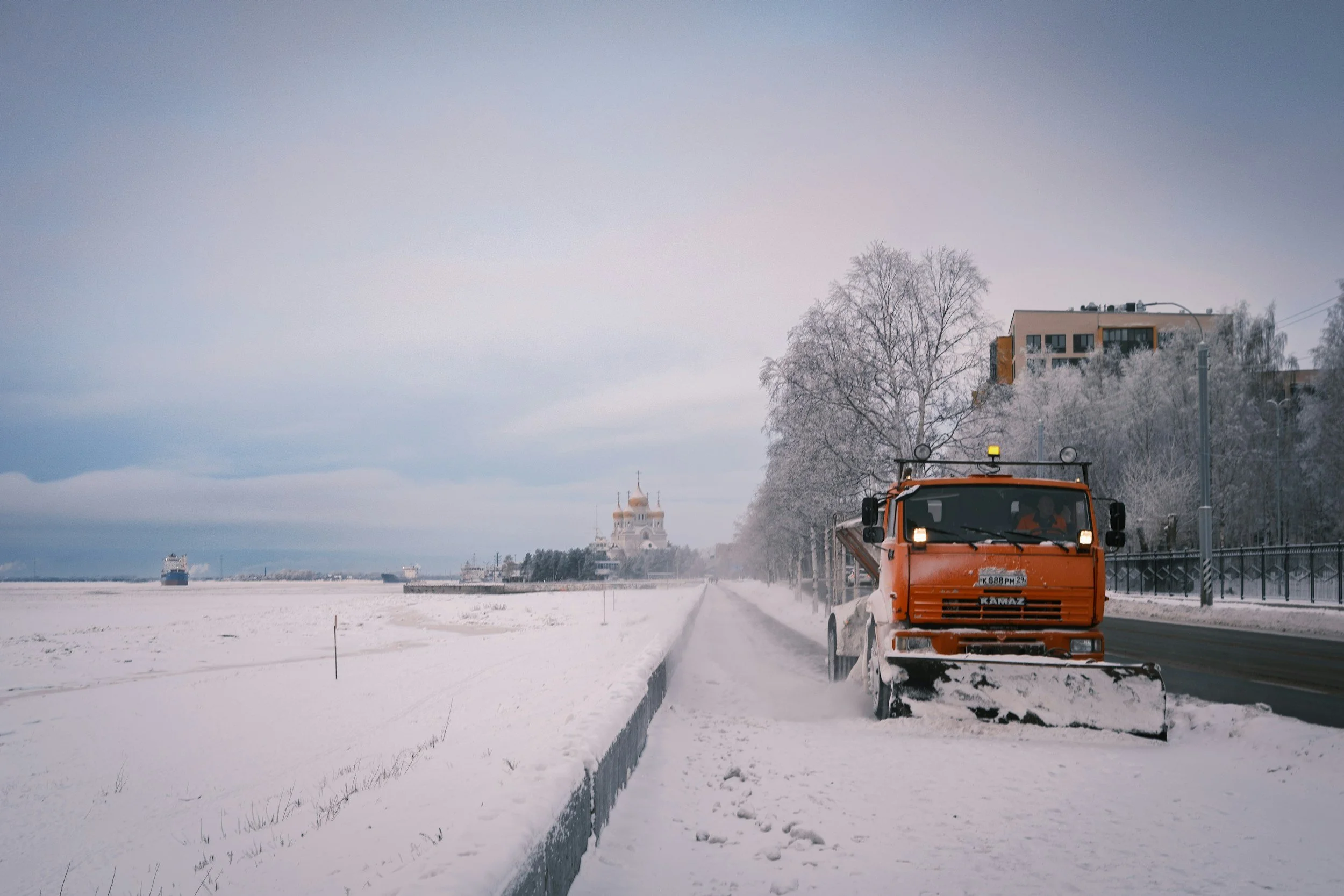 Ein orangefarbener Schneepflug räumt Schnee von einer Straße in einer winterlichen Landschaft mit Bäumen und Gebäuden im Hintergrund.