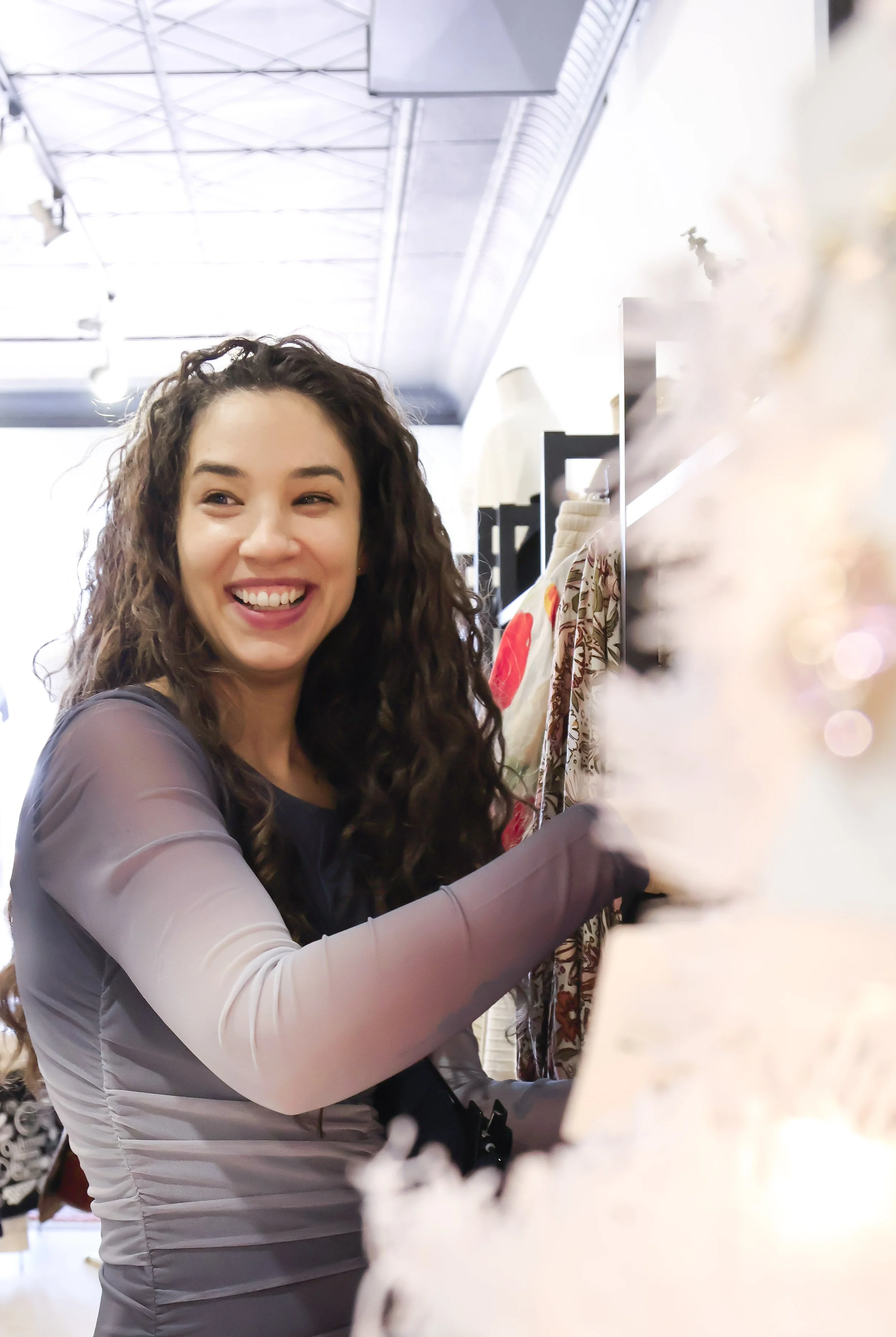 A woman with long curly hair, smiling and shopping in a clothing store. She is reaching toward a rack of clothes.