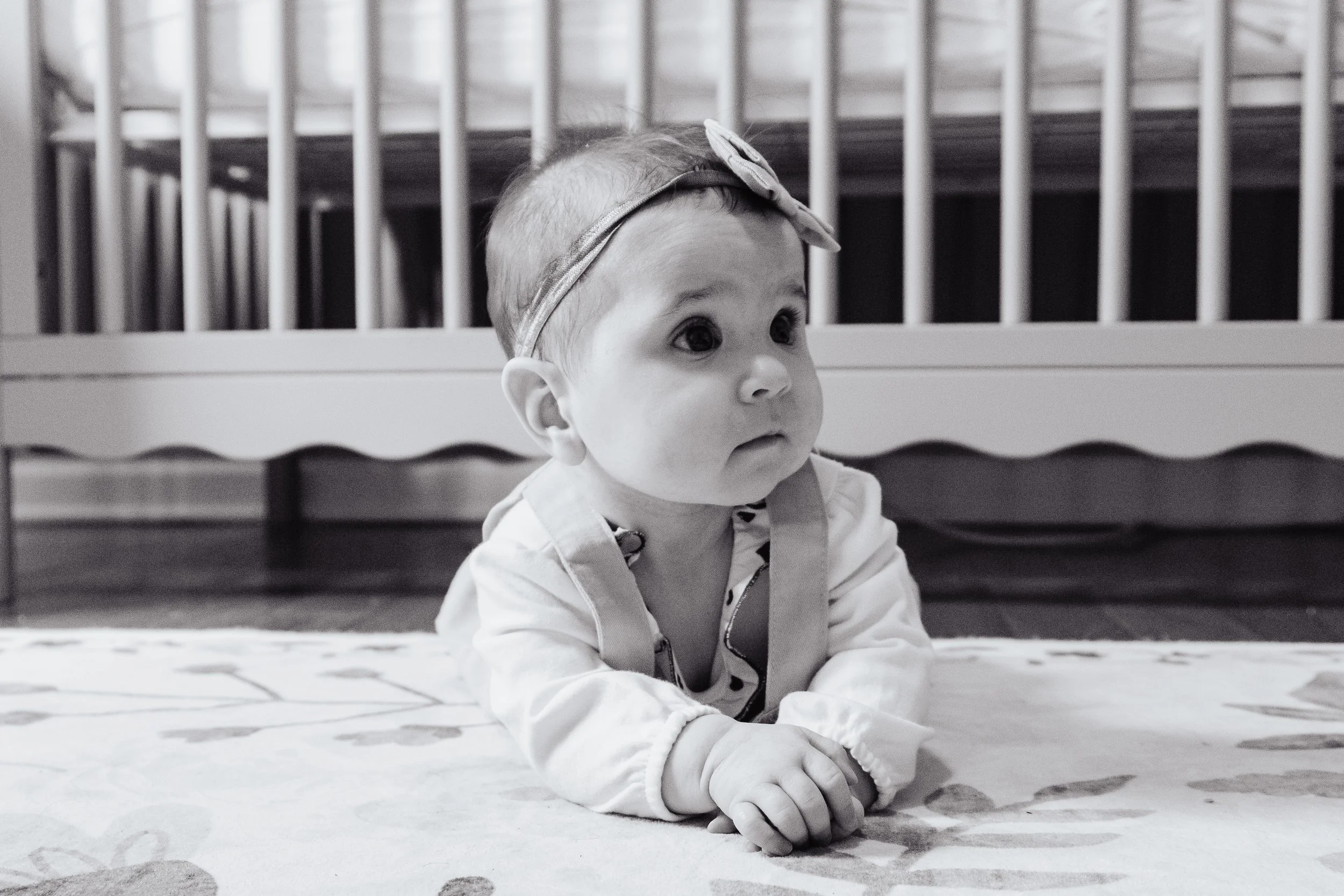 A baby girl with a bow headband lying on her stomach on a patterned rug, with a crib in the background.