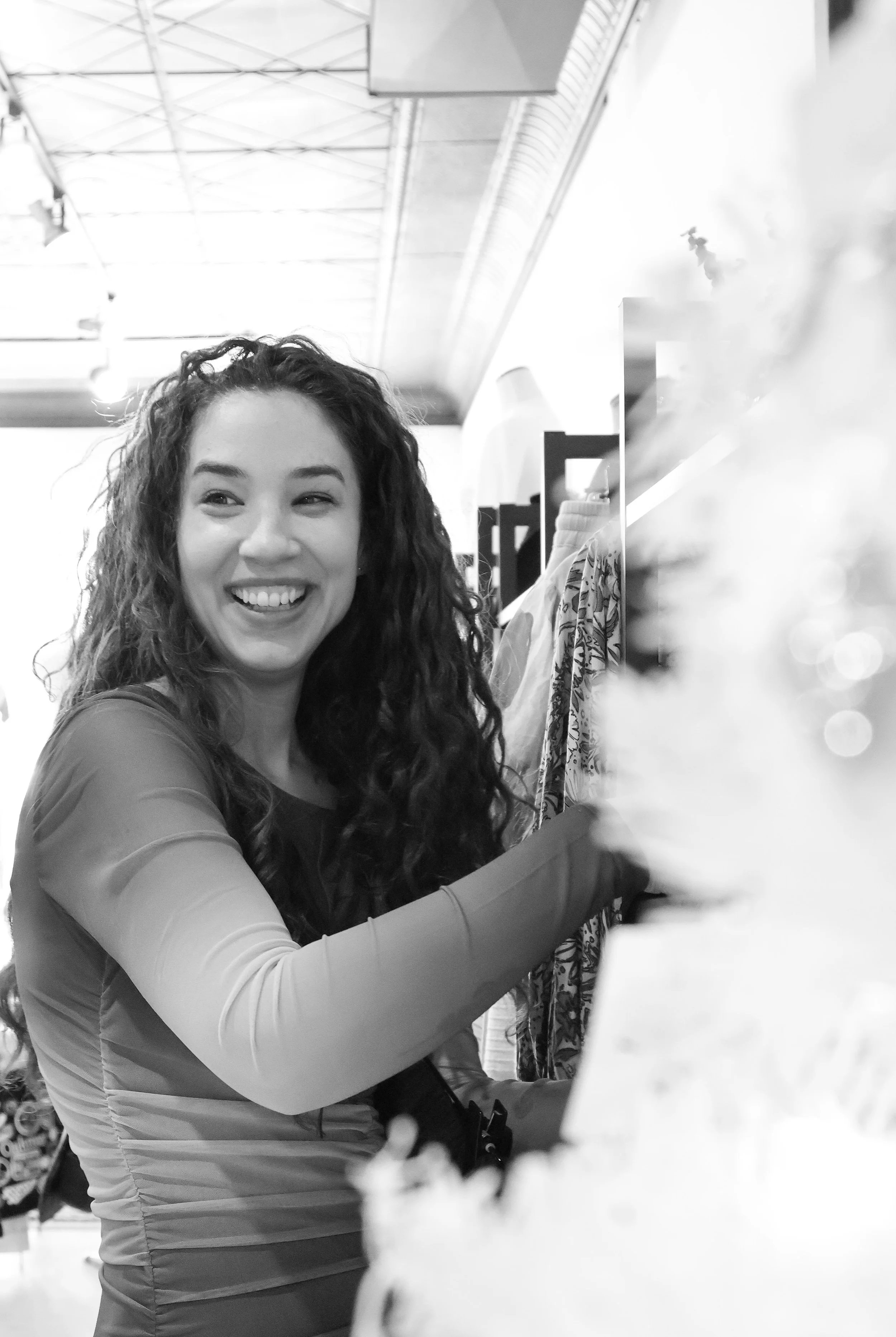 A smiling woman with curly hair shopping in a clothing store, reaching for an item on the rack.