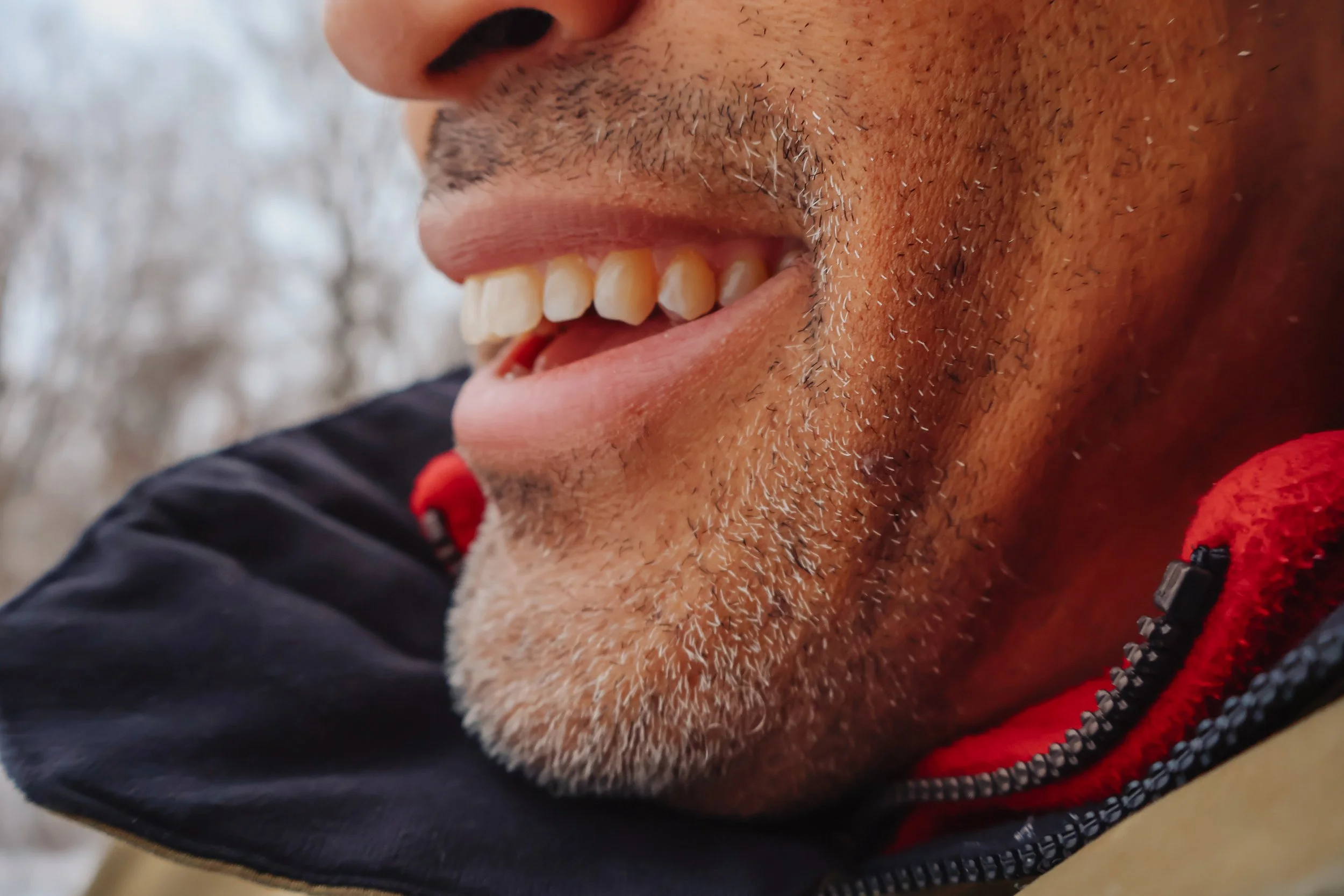 Close-up of a man's face smiling, showing teeth, beard, and mustache, with a blurred background of trees.