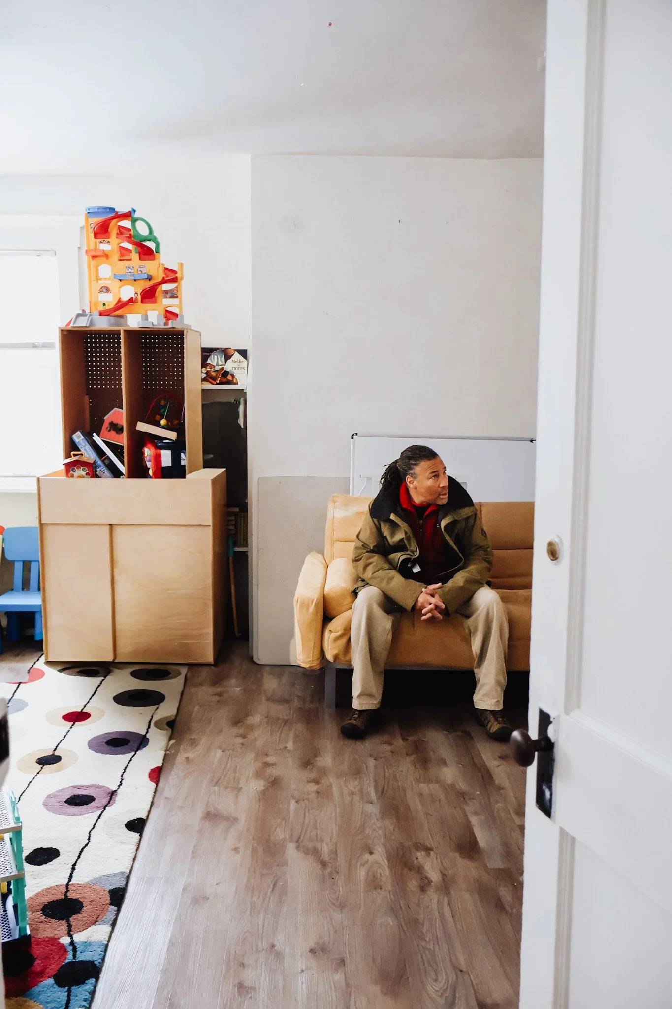 A man sitting on a tan sofa in a room with a white wall, a colorful rug, a wooden storage unit with toys, and a window.