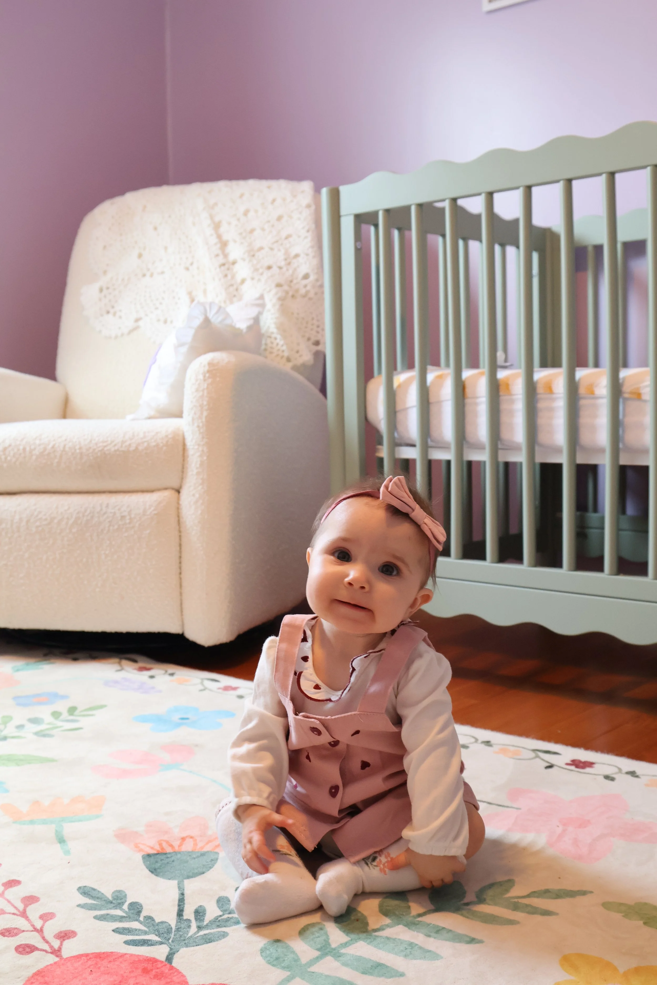 A baby girl with a pink bow headband, sitting on a floral rug in a nursery with a crib and a white armchair in the background.