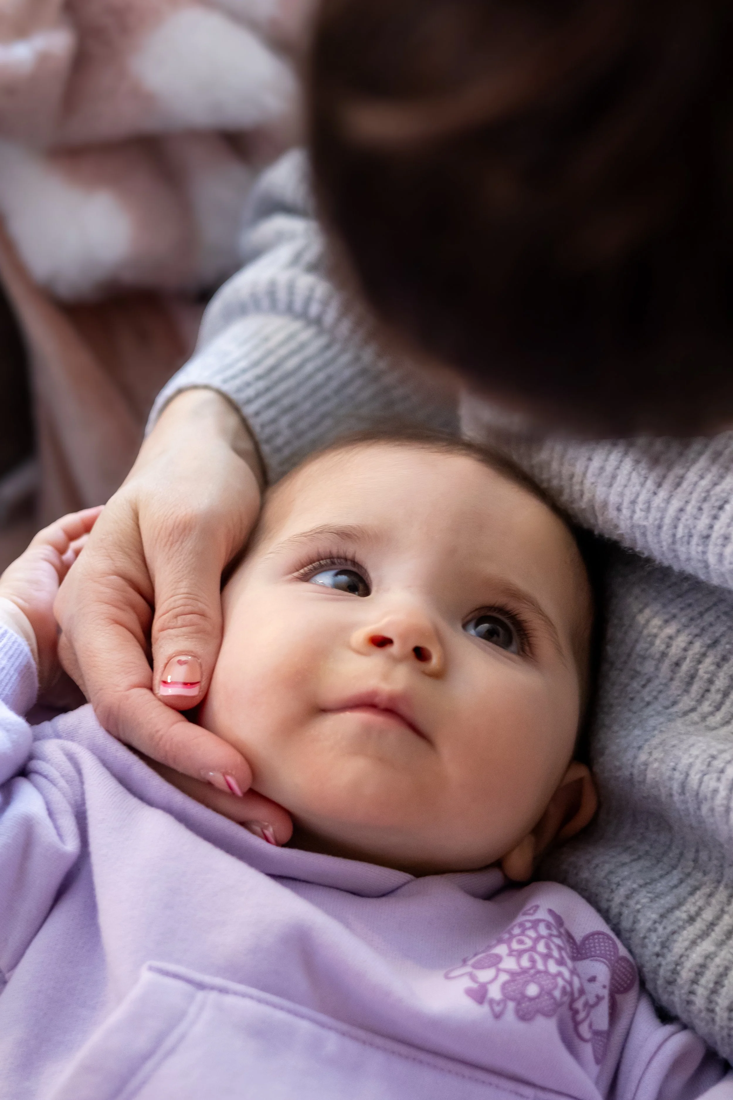 Close-up of a baby lying down with a woman gently holding the baby's face, both looking at each other, soft background with blankets.