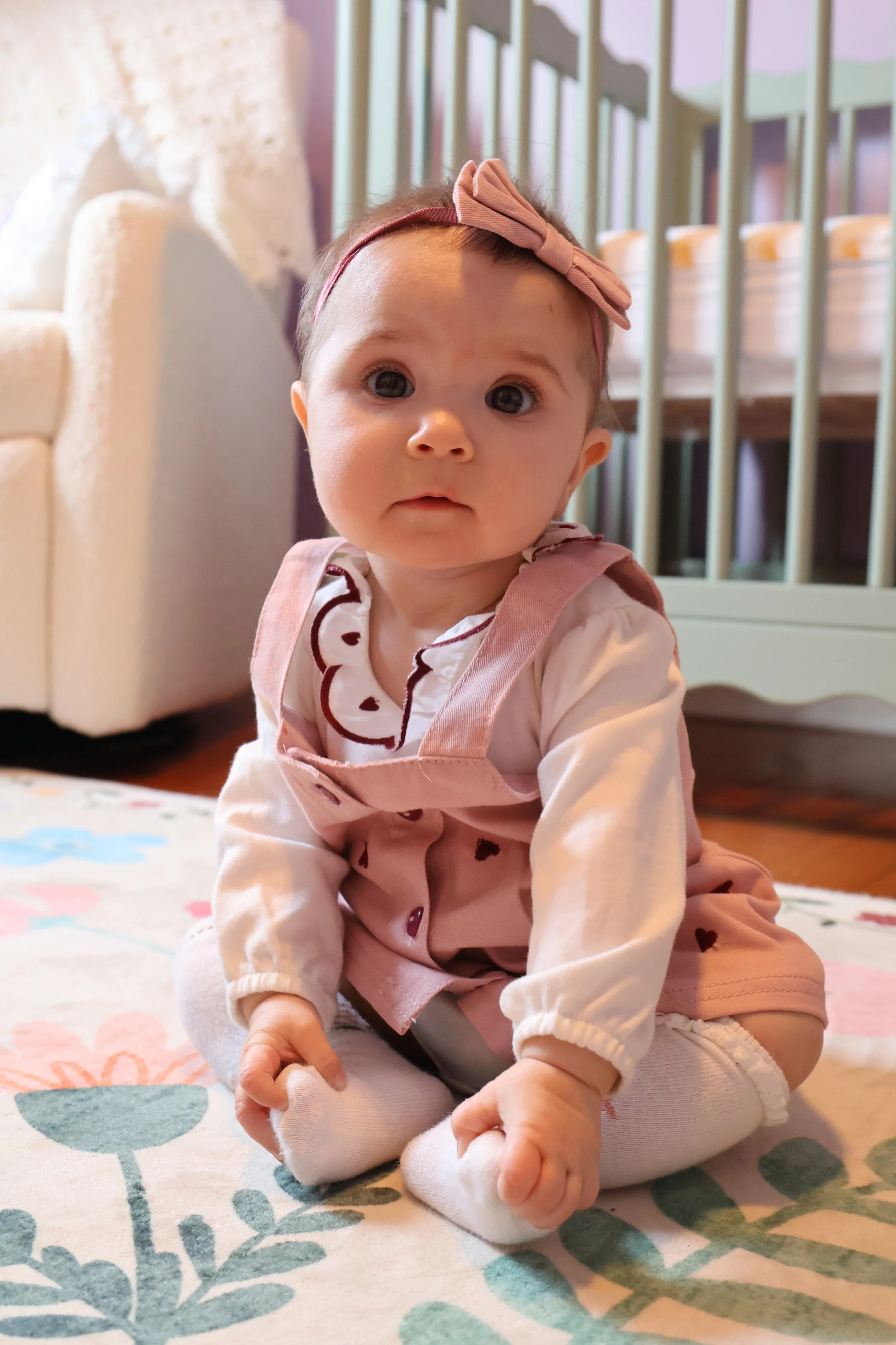A baby girl sitting on a floral carpet in a room with a crib and a sofa in the background.
