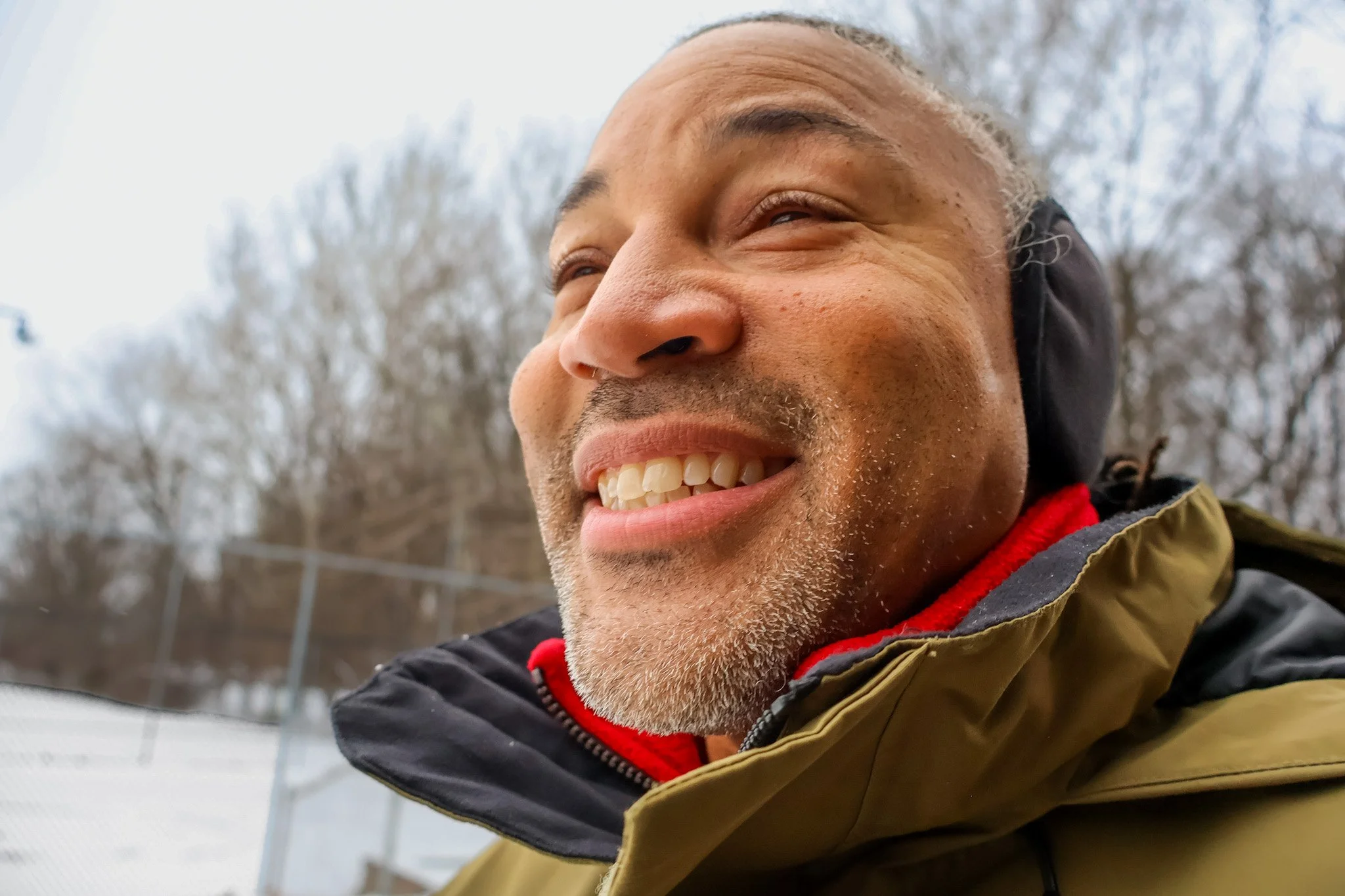 Close-up of a smiling man outdoors, wearing a jacket with a red collar and black hood, with trees in the background.