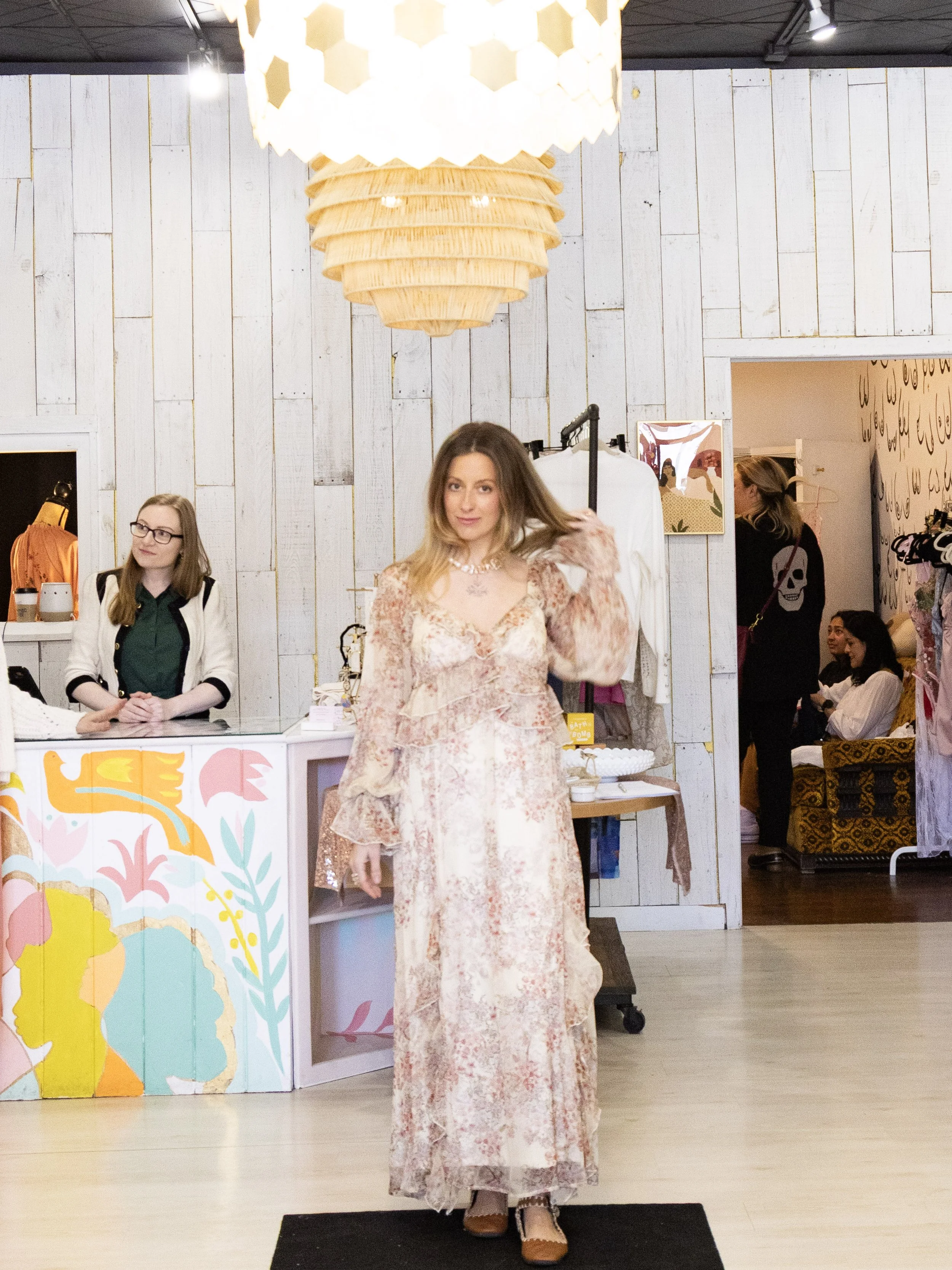A woman in a long, floral dress stands on a black mat inside a boutique or showroom, smiling and looking at the camera. Behind her, there are two women sitting on a yellow and black patterned couch, and another woman browsing clothing.