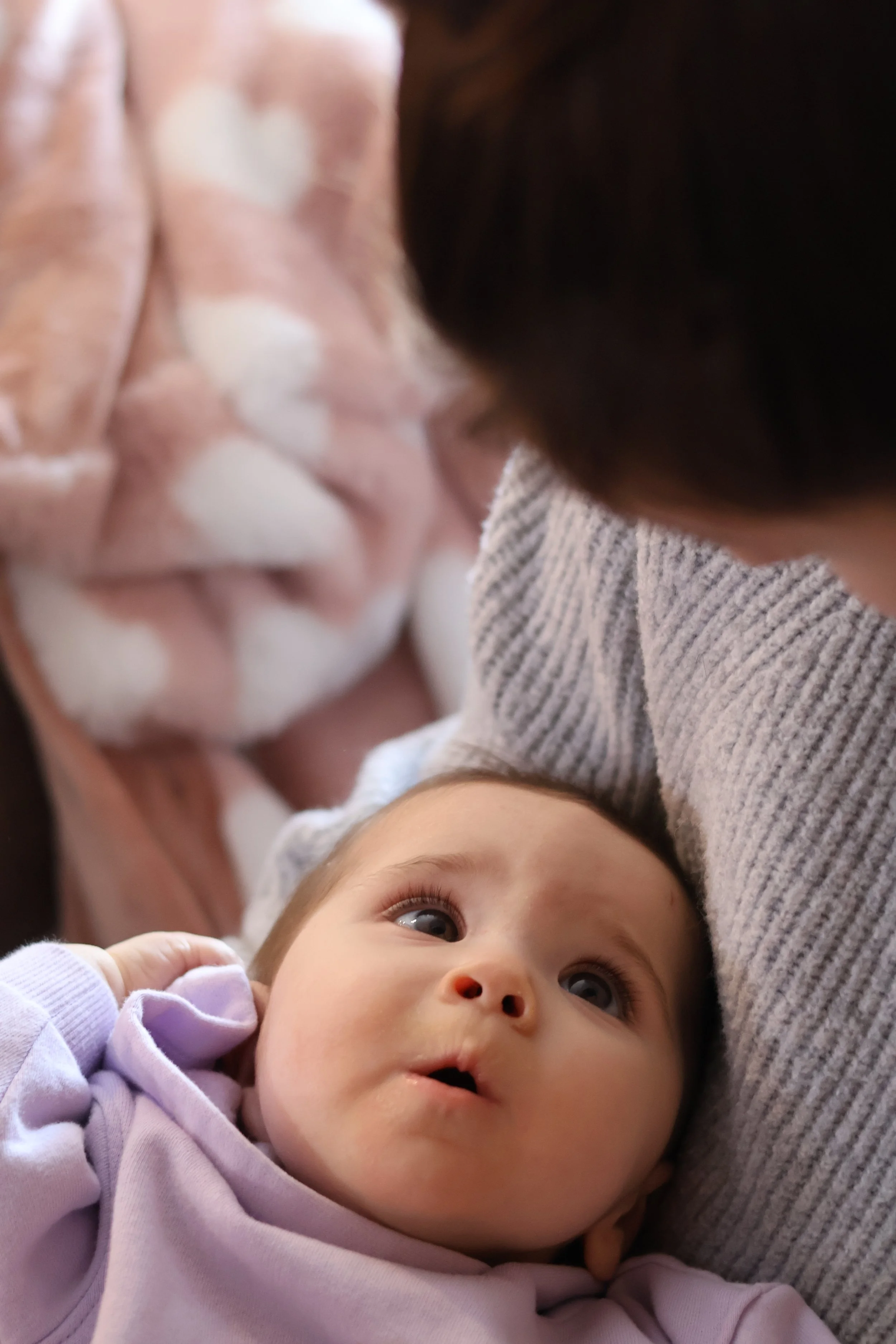 Close-up of a baby lying in someone's arms, with wide eyes and slightly parted lips, looking up at a person wearing a gray sweater, with a blanket in the background.