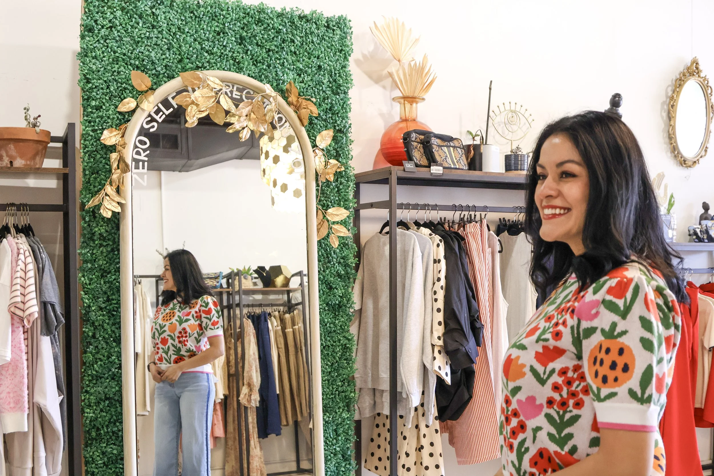 A woman with dark, wavy hair and red lipstick standing in front of a mirror in a clothing store, wearing a colorful floral shirt, and smiling.