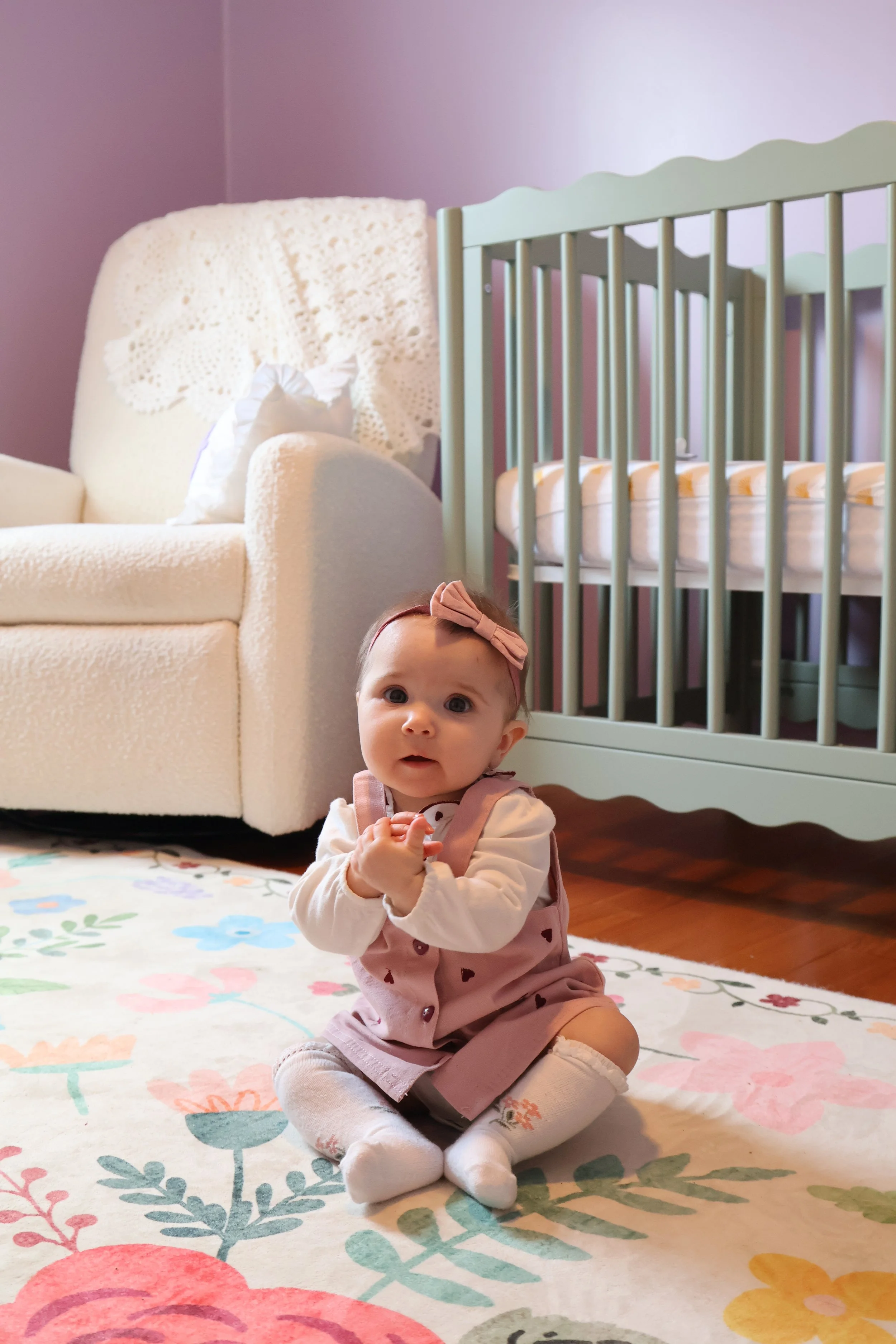 A baby girl sitting on a floral rug in a living room with a white sofa and a green crib in the background.