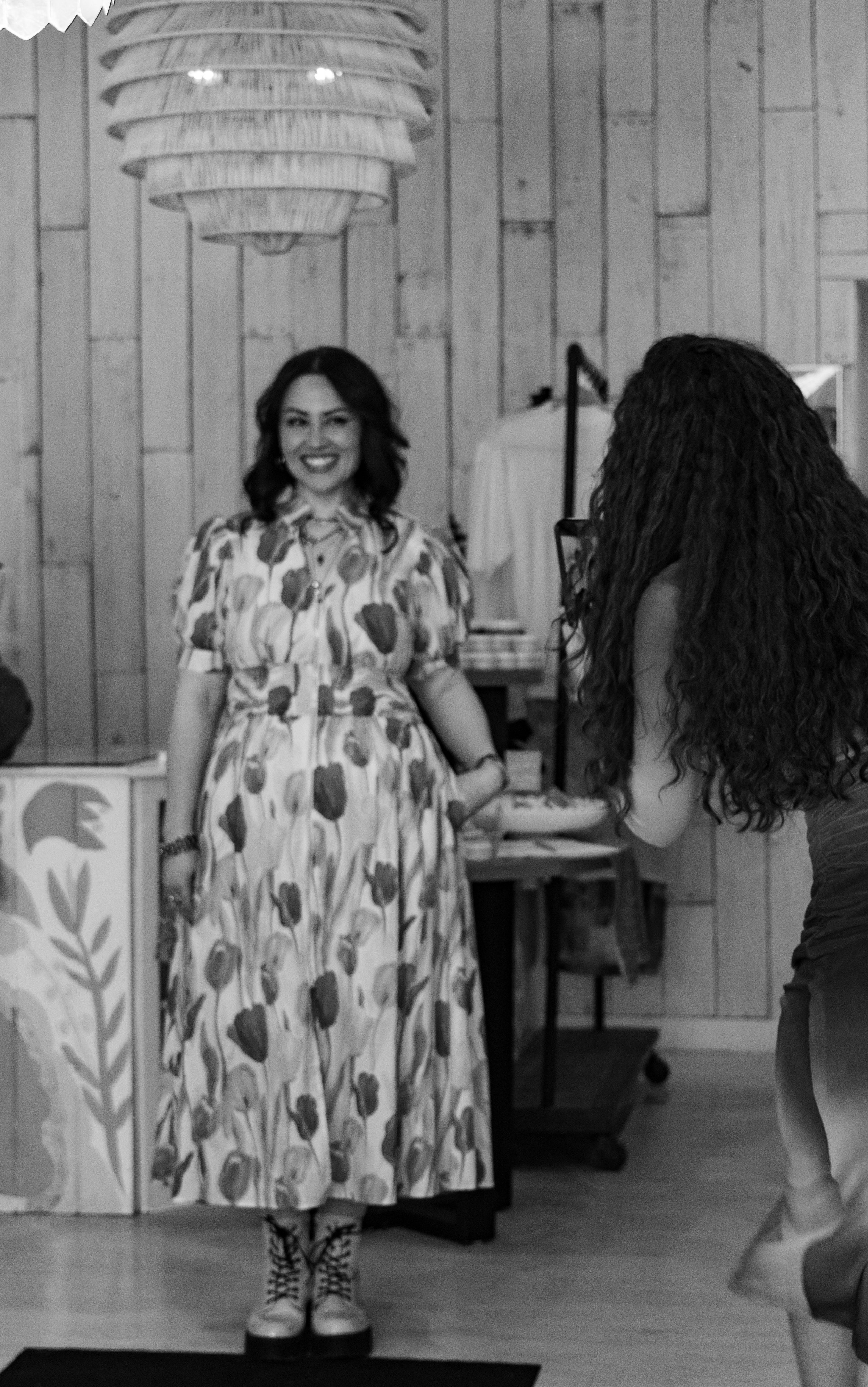 A woman in a floral dress standing and smiling, with someone taking her photo in a wooden interior space.