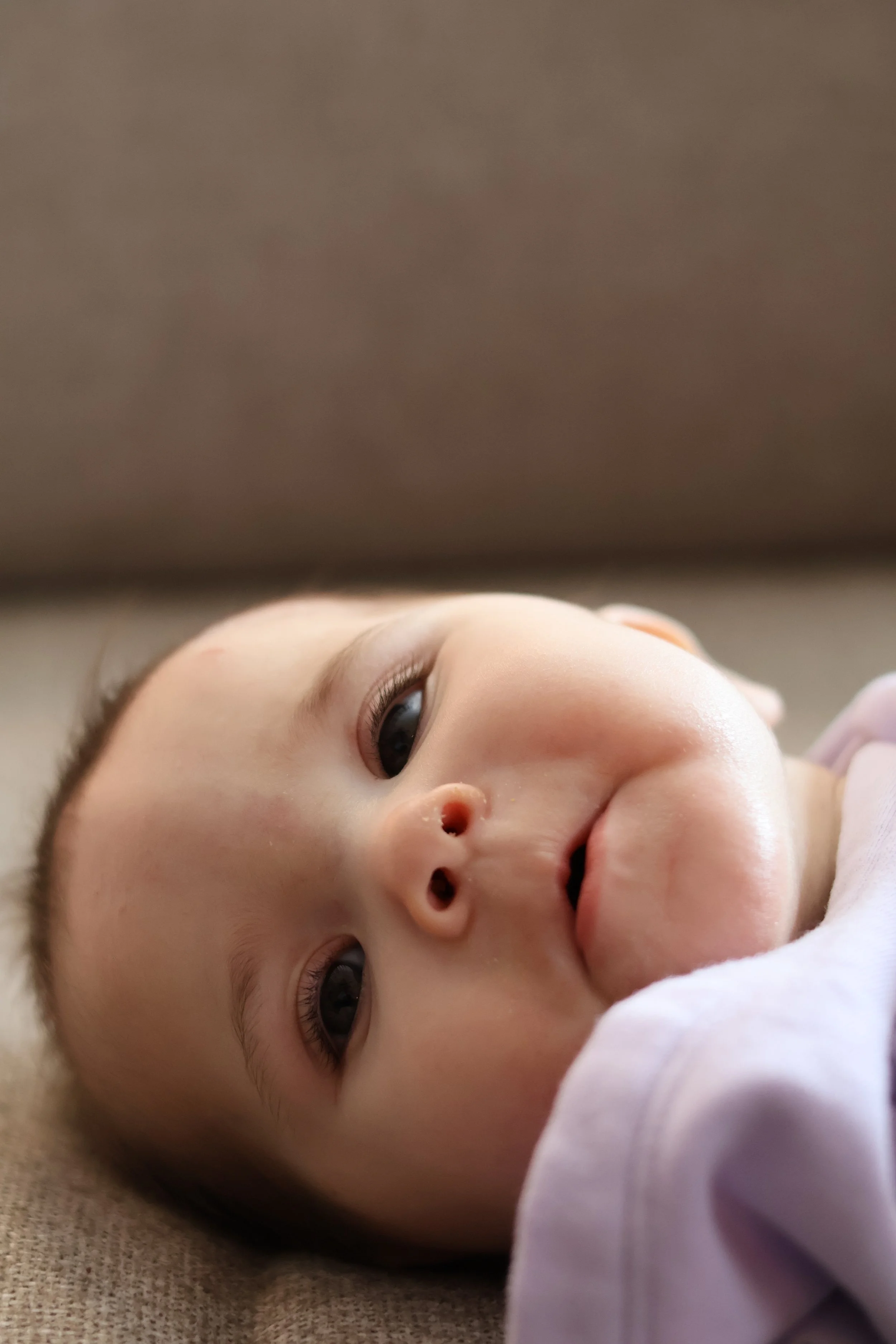 Close-up of a baby lying on a soft surface, looking up with wide eyes, wearing a light-colored outfit.