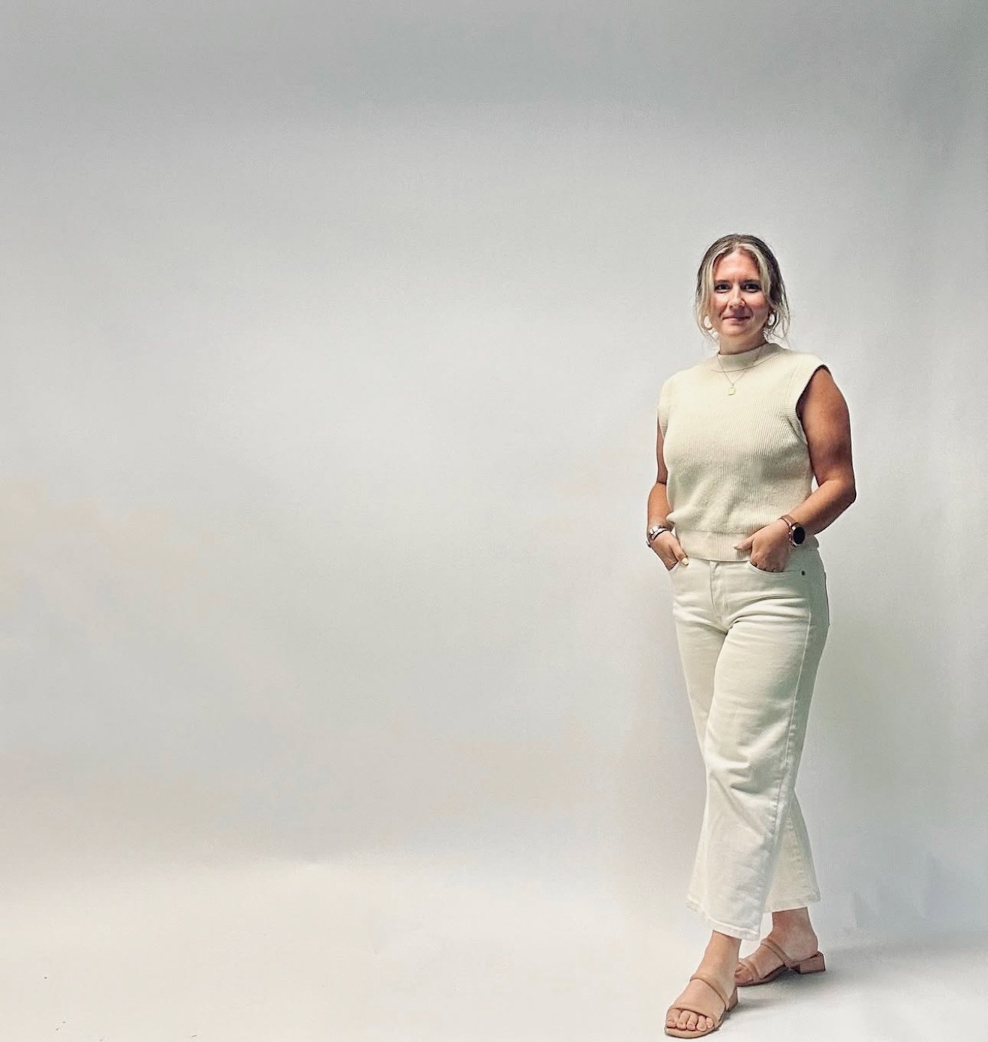 A woman with blonde hair is standing in front of a plain white wall, wearing a sleeveless beige top, off-white pants, and beige heeled sandals.