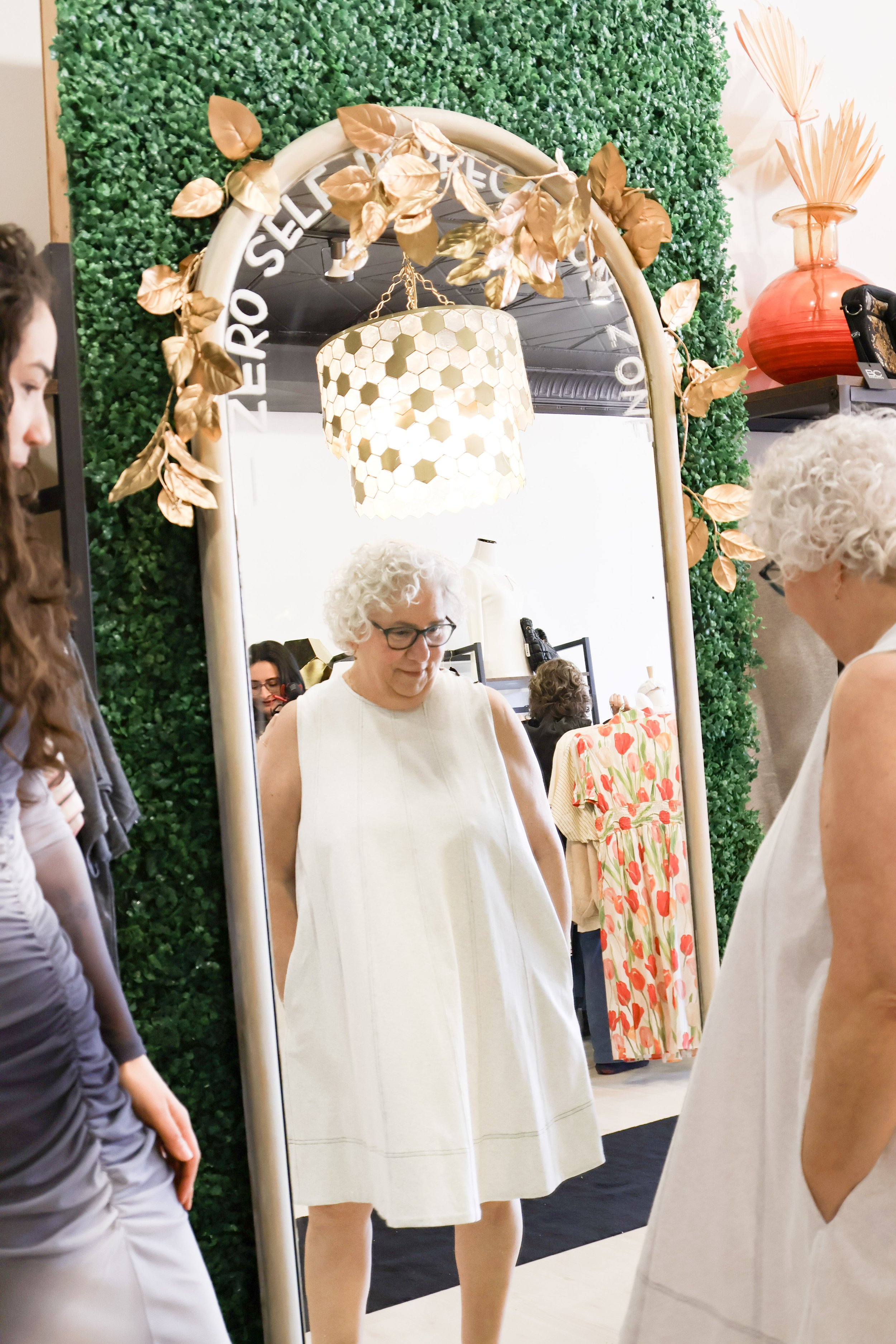 An elderly woman with curly white hair and glasses wearing a sleeveless white dress, standing in front of a mirror in a boutique, trying on a similar white dress, surrounded by other women, with a green leafy wall, a hanging hexagonal lampshade, and 