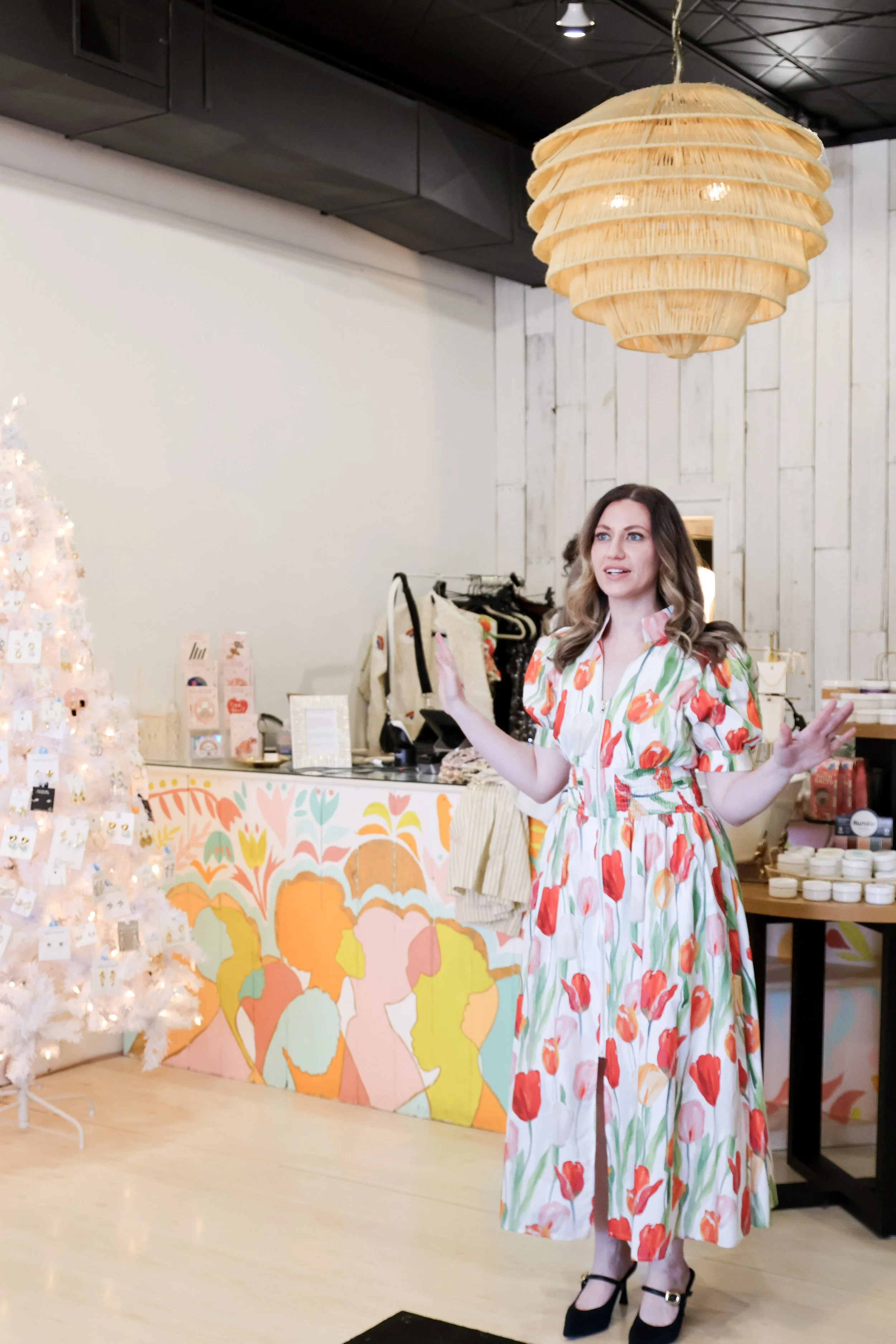 Woman in a floral dress speaking in a decorated retail space with a white Christmas tree and colorful artwork on the wall.