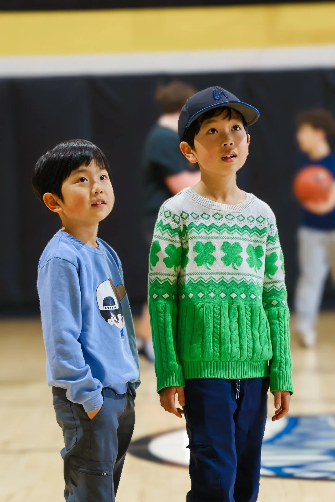 Two young boys standing in a gymnasium, watching something off-camera, with a person holding a basketball in the background.