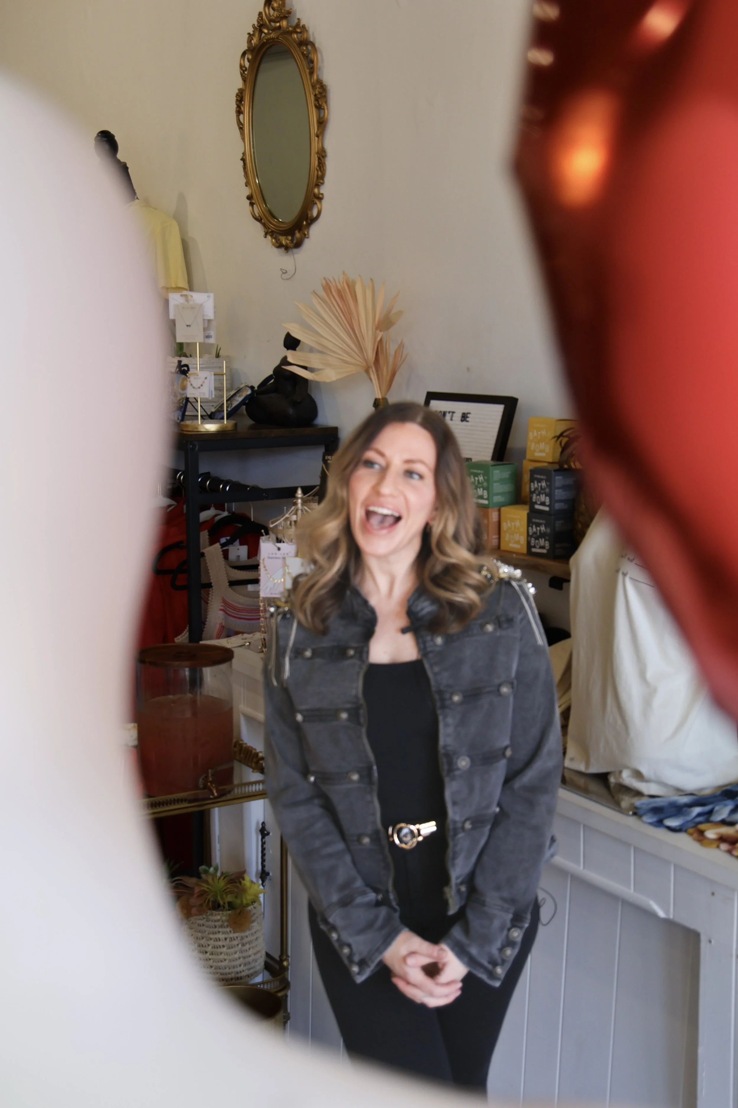A woman with wavy hair smiling and standing in a store, viewed through the colorful foreground of other people's clothing or accessories.
