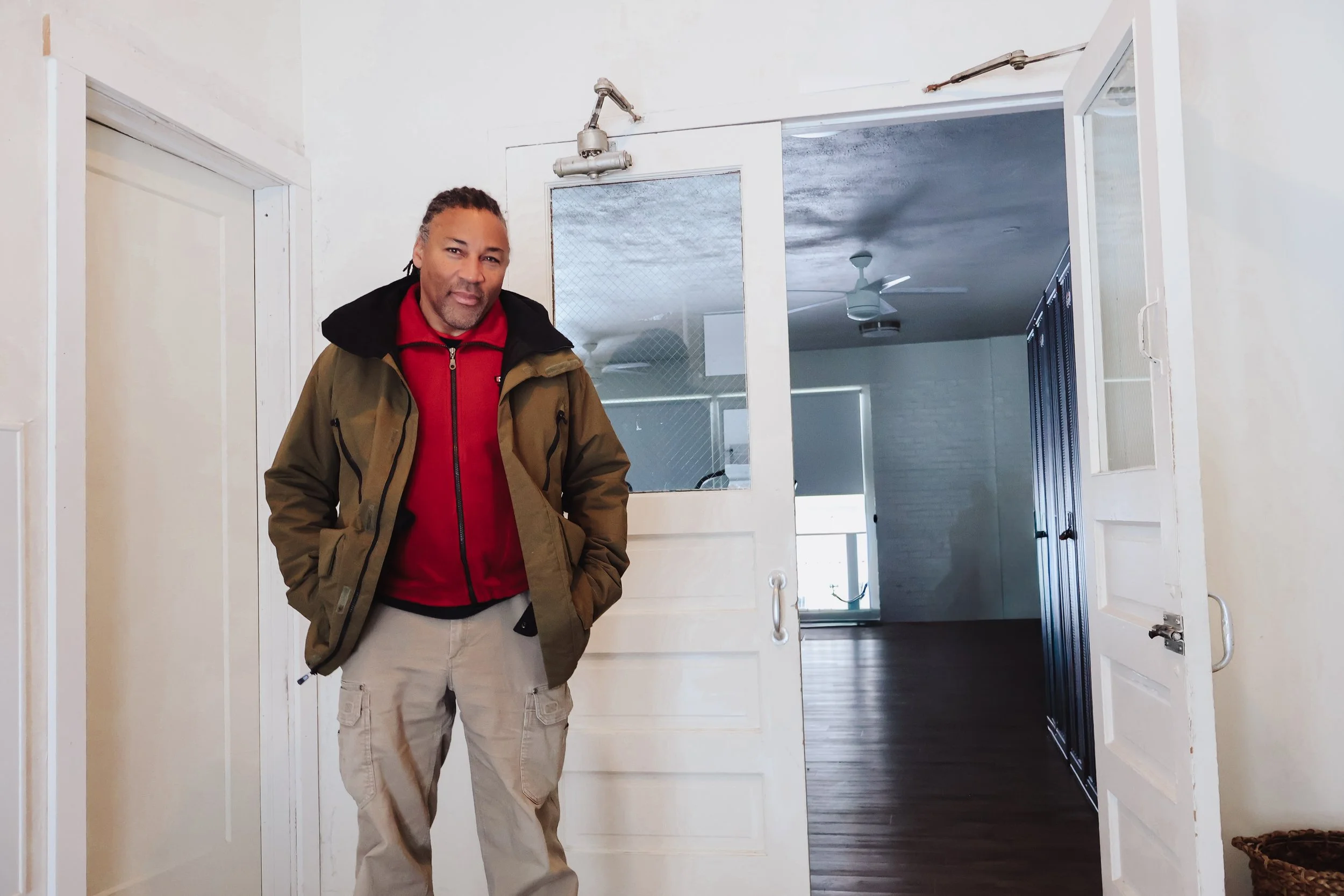 A man with long dreadlocks, wearing a red jacket and an olive green parka, stands in front of a white door in a house.