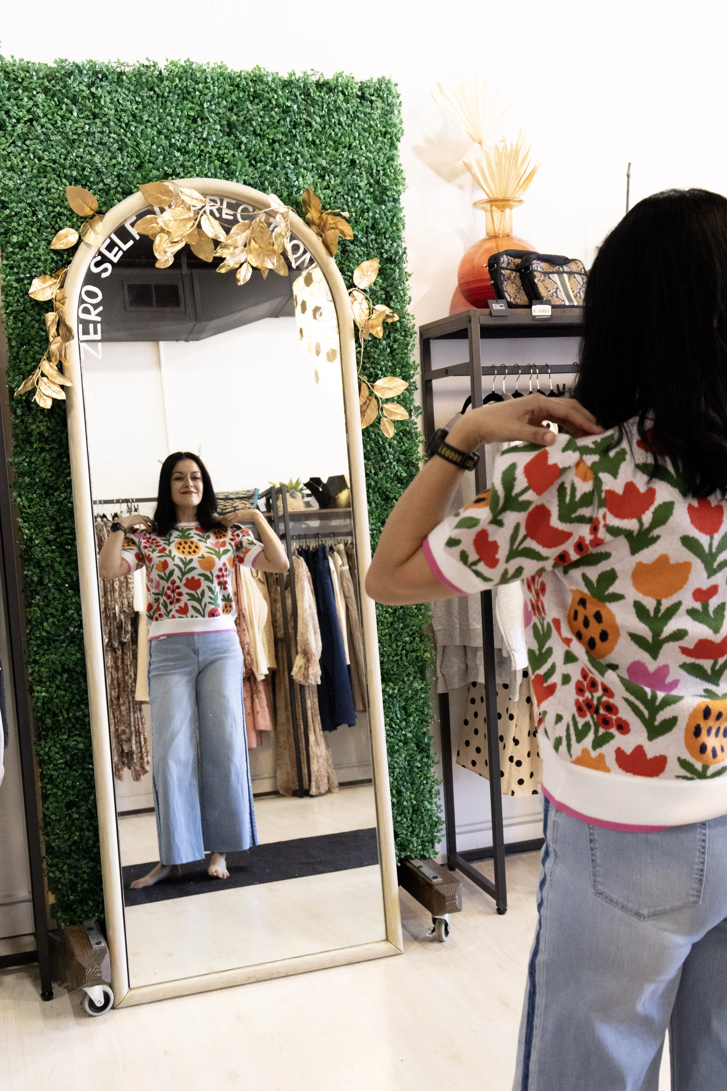 Woman trying on a colorful floral sweater and wide-leg jeans looks at herself in a large decorative mirror inside a boutique. The mirror is framed with golden leaves and greenery, with clothing racks and decorative items visible in the background.