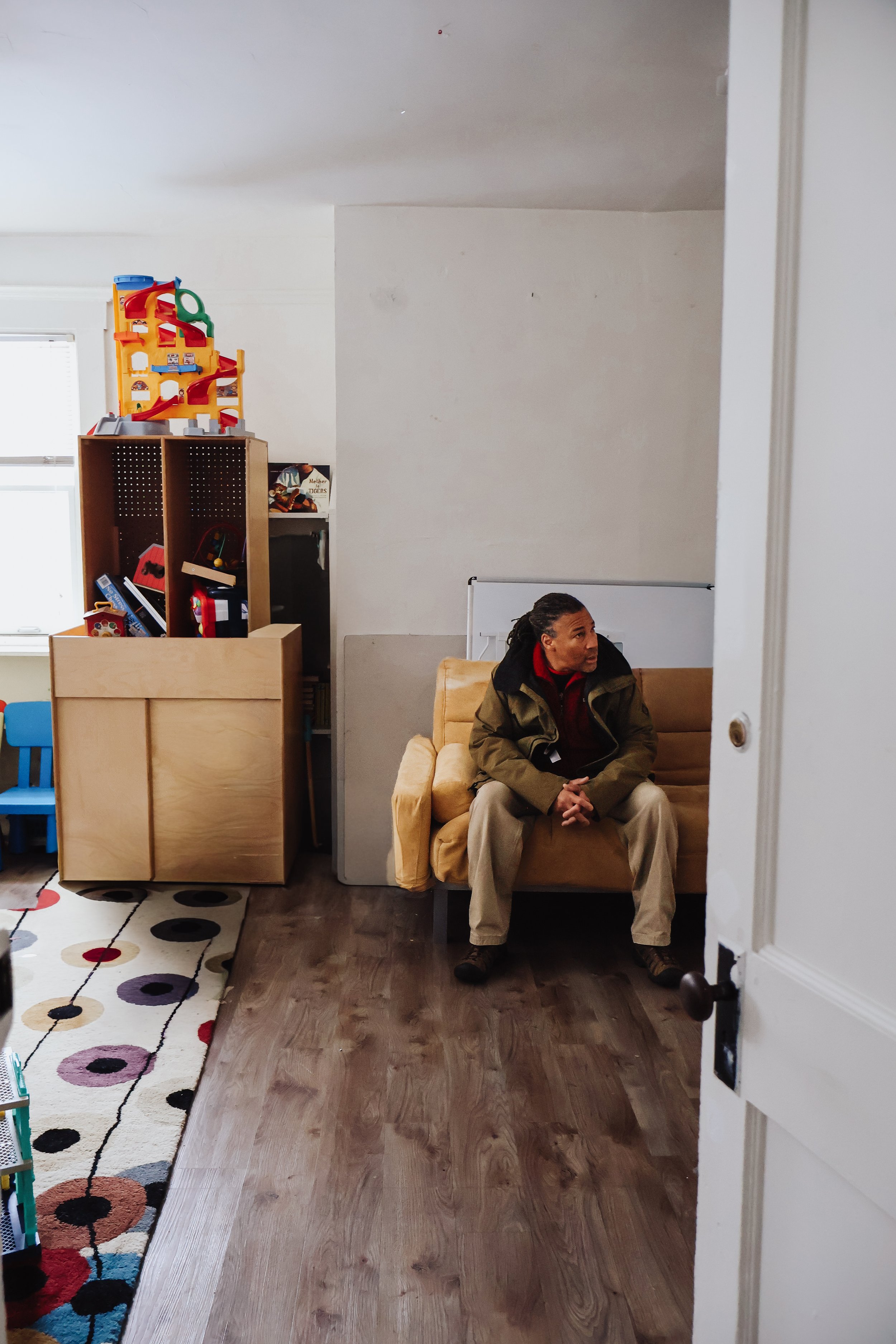 A man sitting on a yellow couch in a room with wooden floors, a white wall, a window, a colorful toy storage unit, and a partially visible open door.