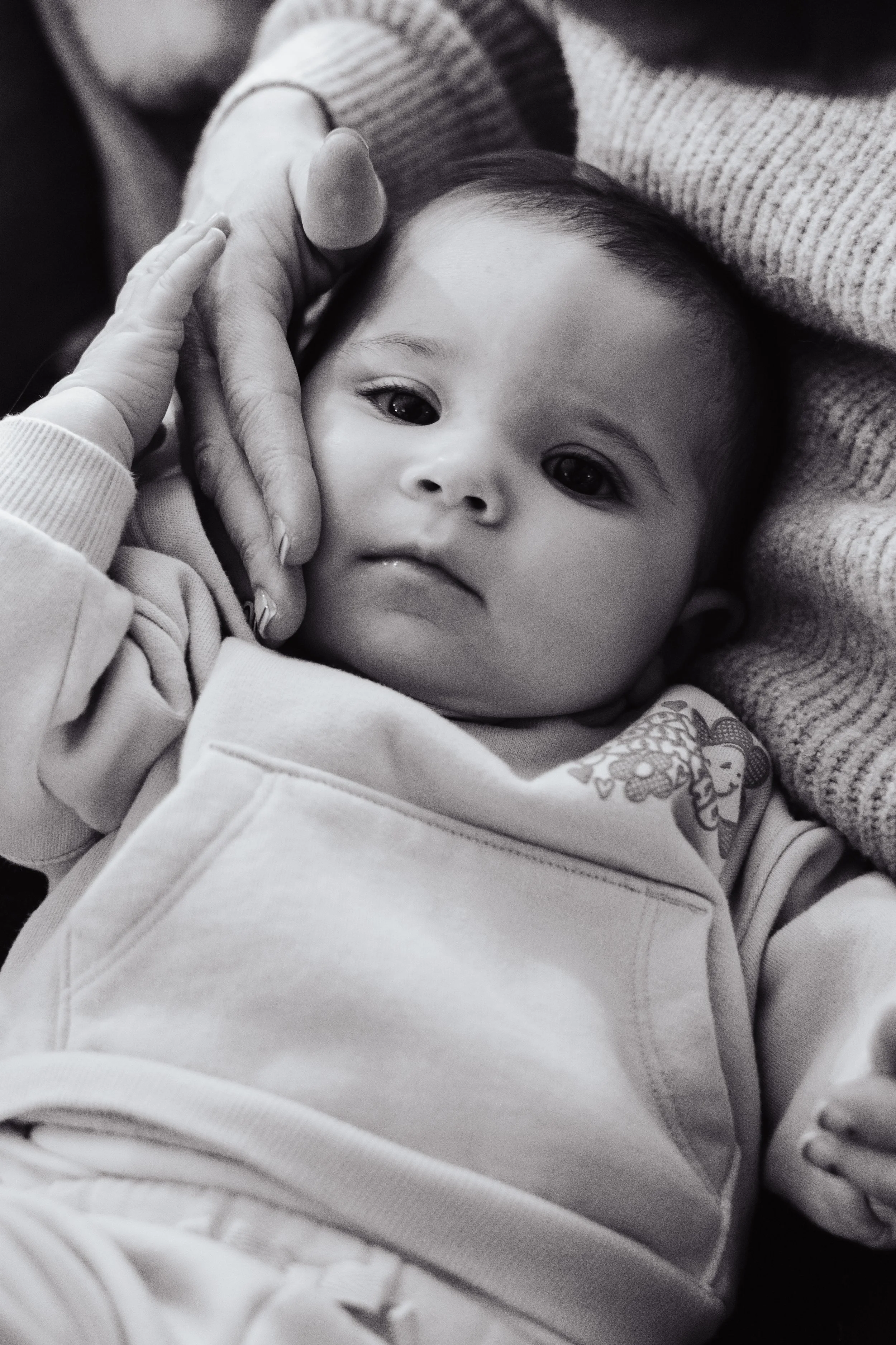 A black and white photo of a baby lying down, being gently held by an adult's hand and face. The baby has big, dark eyes and is dressed in a hoodie with a cartoon character on it.