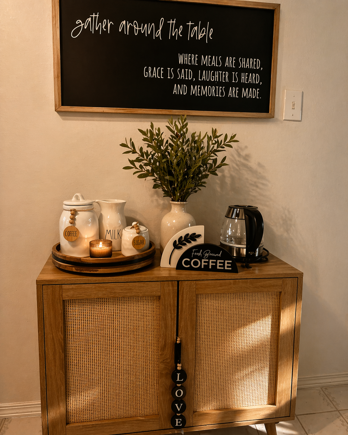 A wooden sideboard with white jars labeled coffee, milk, and sugar, a potted plant, a small candle, and a glass coffee maker. Above, a black chalkboard sign reads 'gather around the table' with a quote about sharing meals, laughter, and memories.