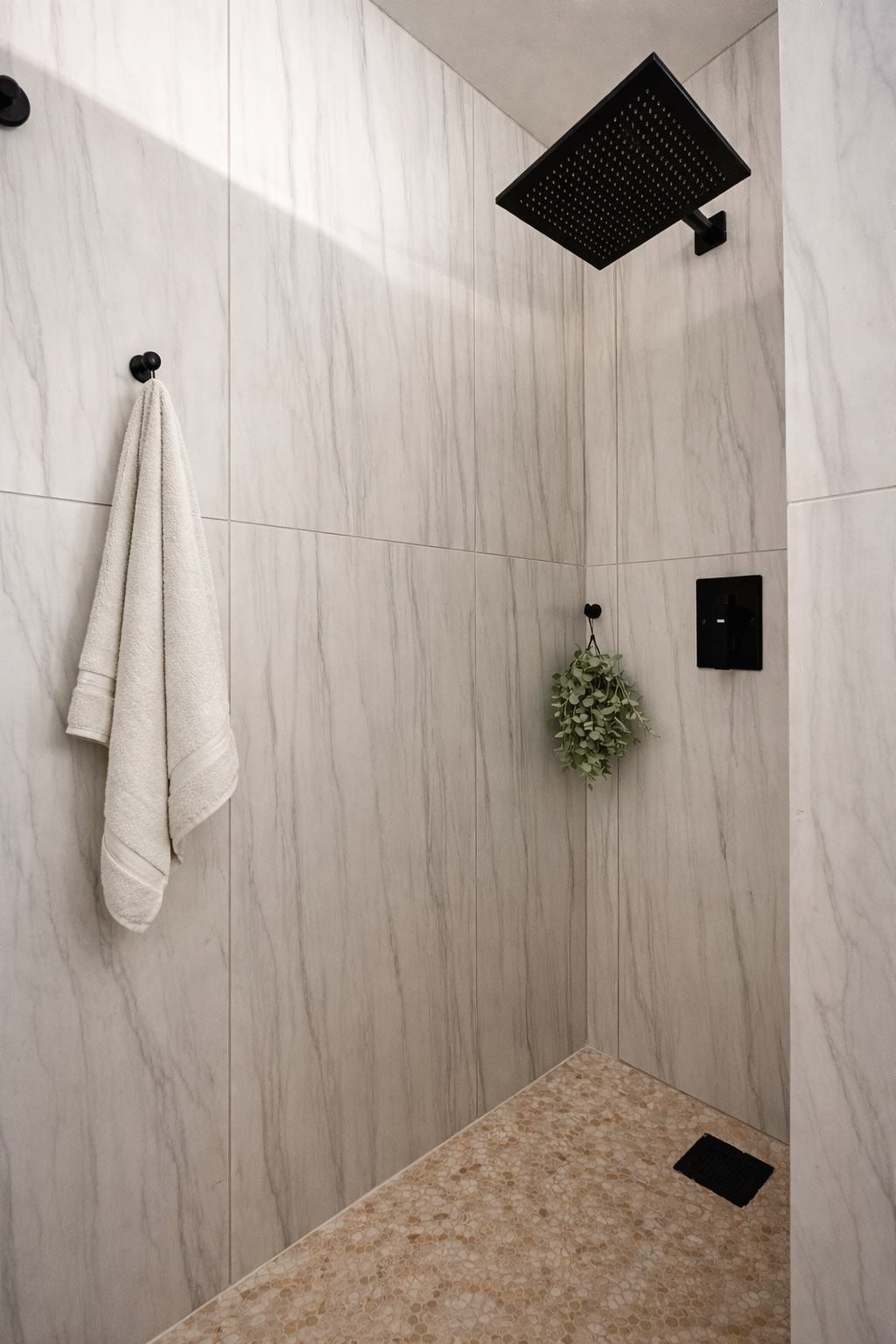 Minimalist shower with a black rain showerhead, a hanging towel, a small hanging plant, and light-colored tiled walls.