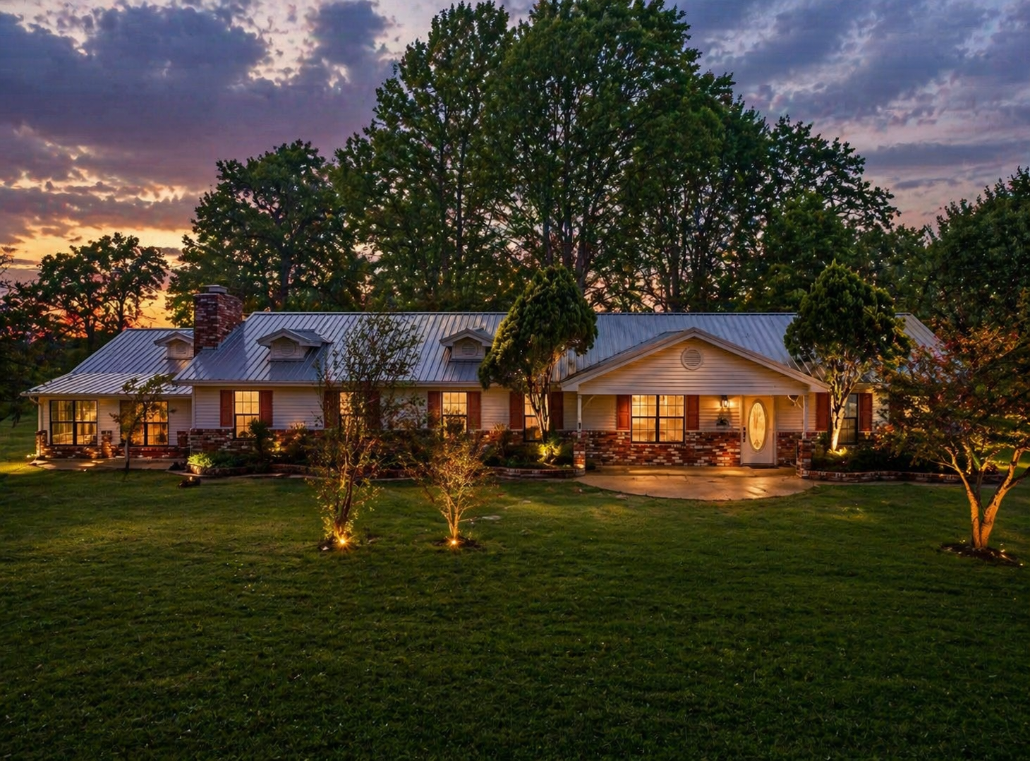 A well-lit house with a metal roof, brick and siding exterior, surrounded by trees and a landscaped lawn during sunset.