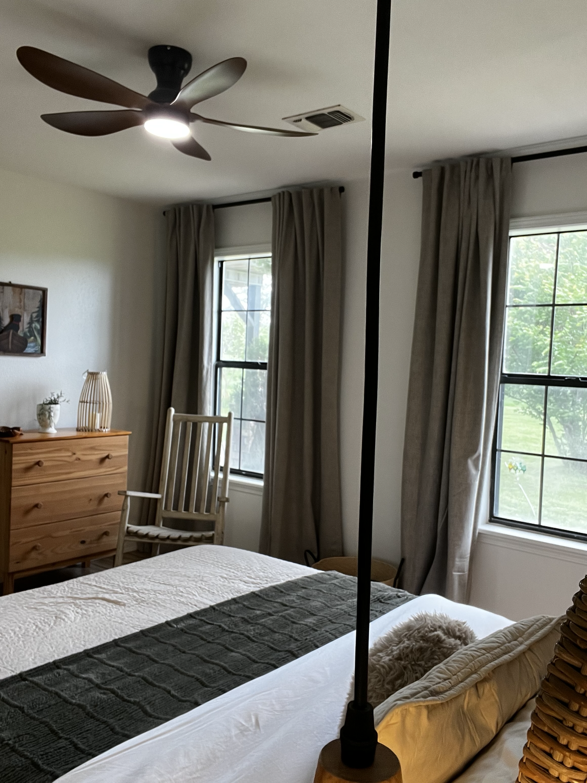 View of a bedroom with two windows, beige curtains, a hanging ceiling fan, a wooden dresser with decorative items, a wooden chair, and a bed with a black throw and a small fluffy dog on it.