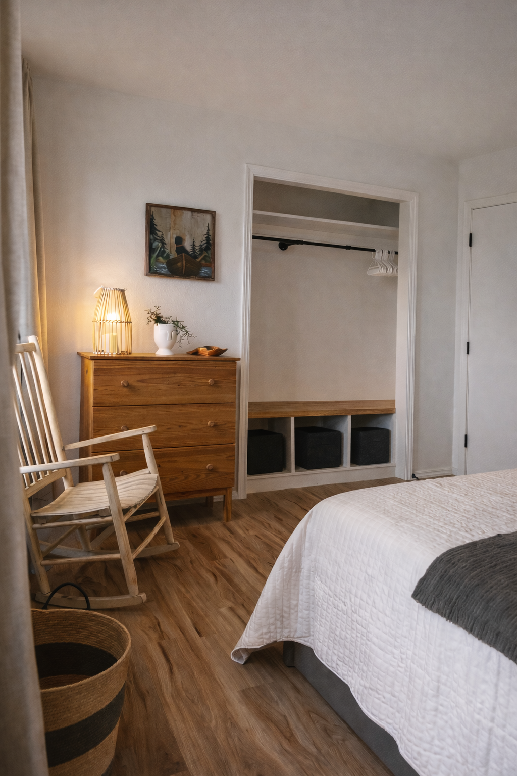 Cozy bedroom with a wooden dresser, a lamp, a plant in a white vase, a framed nature painting, a wooden rocking chair, and a sleeping area with built-in storage and black ottomans.