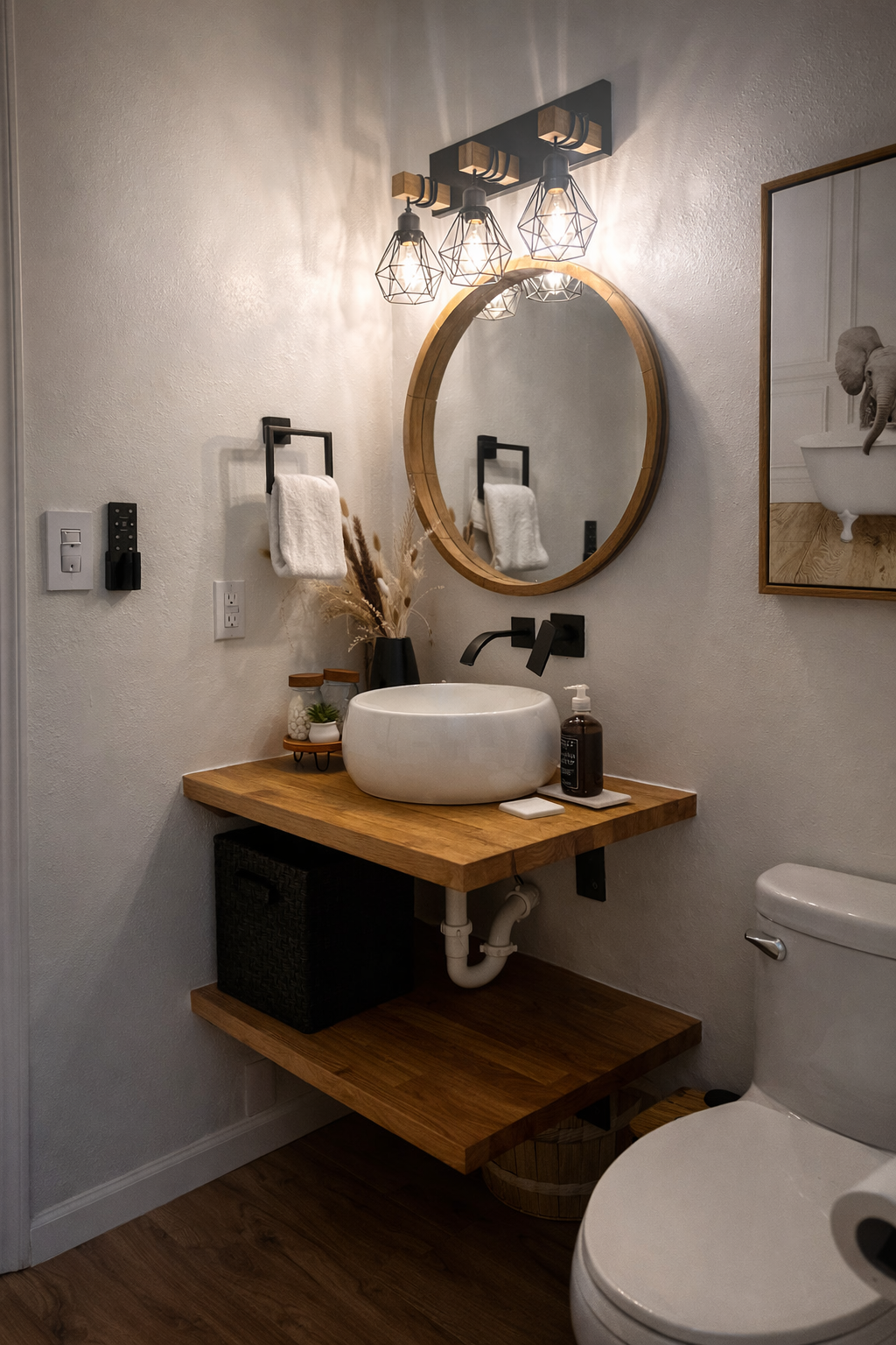 A modern bathroom with a round mirror, industrial-style light fixture, and a wooden vanity with a vessel sink. Decor includes a black vase with dried plants, small containers, and artwork of an elephant. A towel is hanging on a black towel ring next 